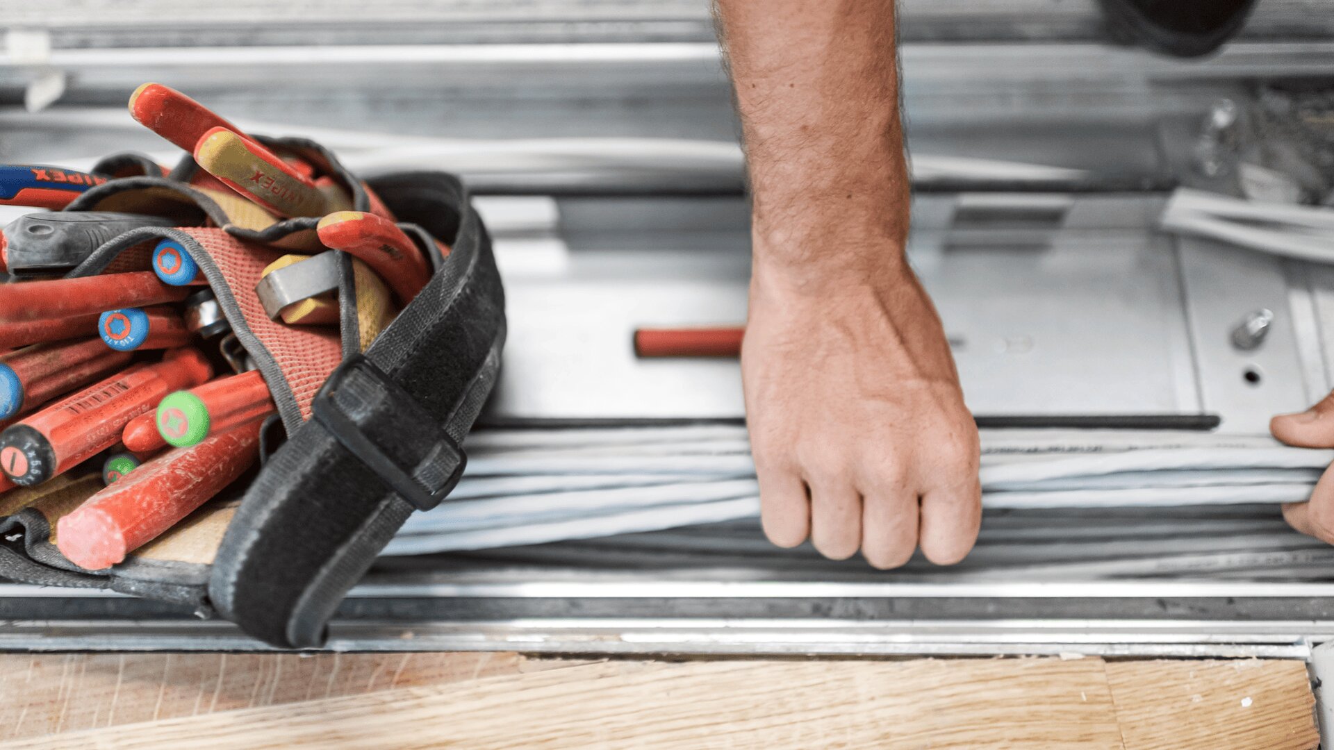 Die Hand einer Person ordnet elektrische Kabel in einem Metallrohr, während eine Werkzeugtasche mit isolierten Schraubenziehern und Zangen in der Nähe auf einem Holzboden liegt.