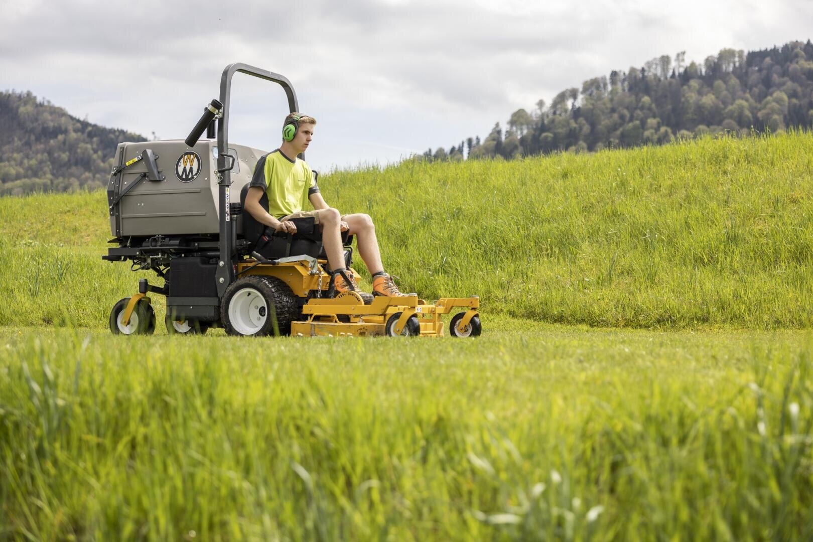 Eine Person mit Kopfhörern bedient einen gelben Aufsitzrasenmäher und mäht Gras auf einem großen, offenen, grünen Feld mit Hügeln und Bäumen im Hintergrund.