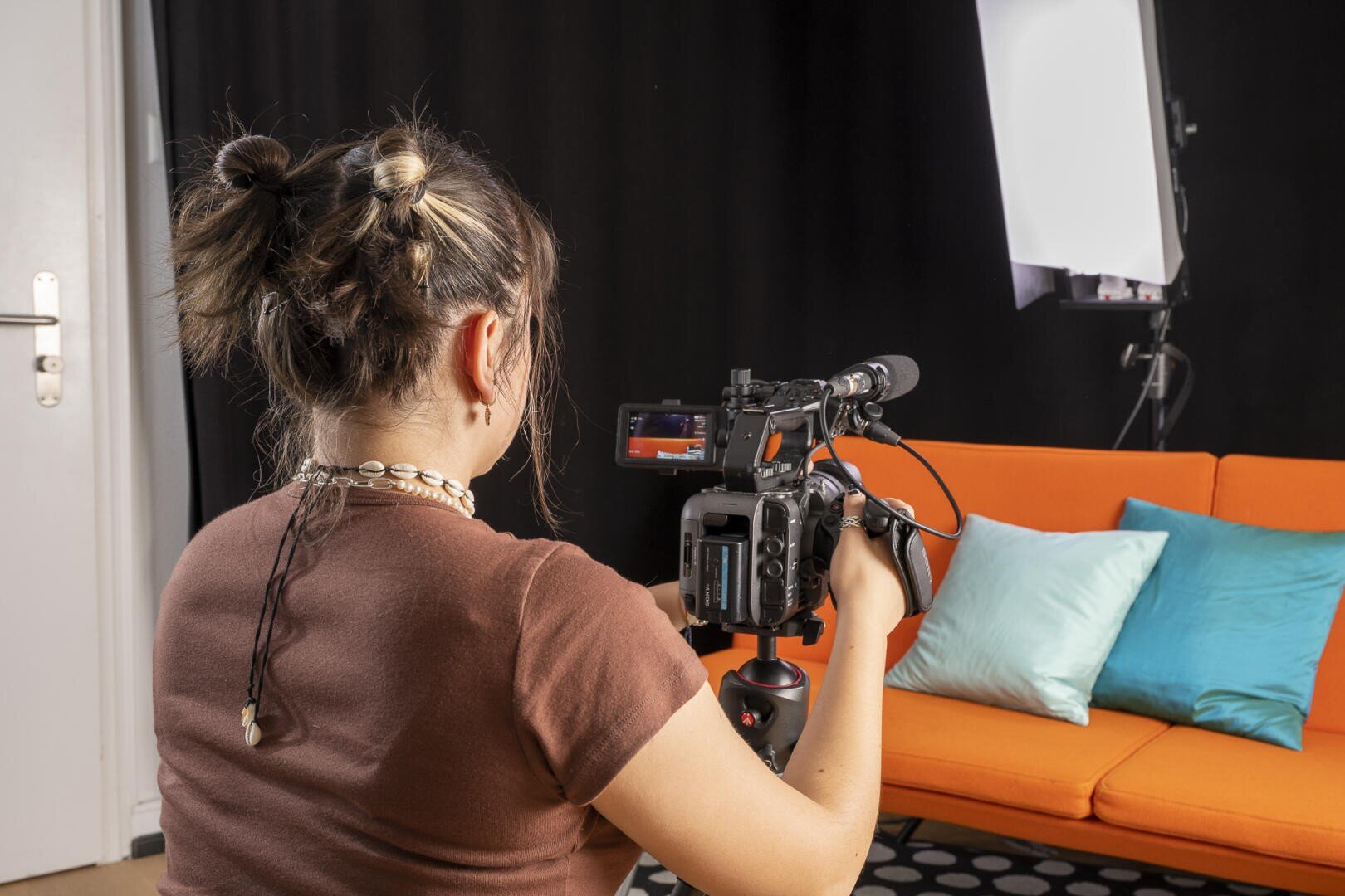 Eine Person mit braunem Haar in einem Dutt filmt ein orangefarbenes Sofa mit blauen und weißen Kissen mit einer professionellen Videokamera in einem Studio mit schwarzem Hintergrund und Beleuchtungsausrüstung.
