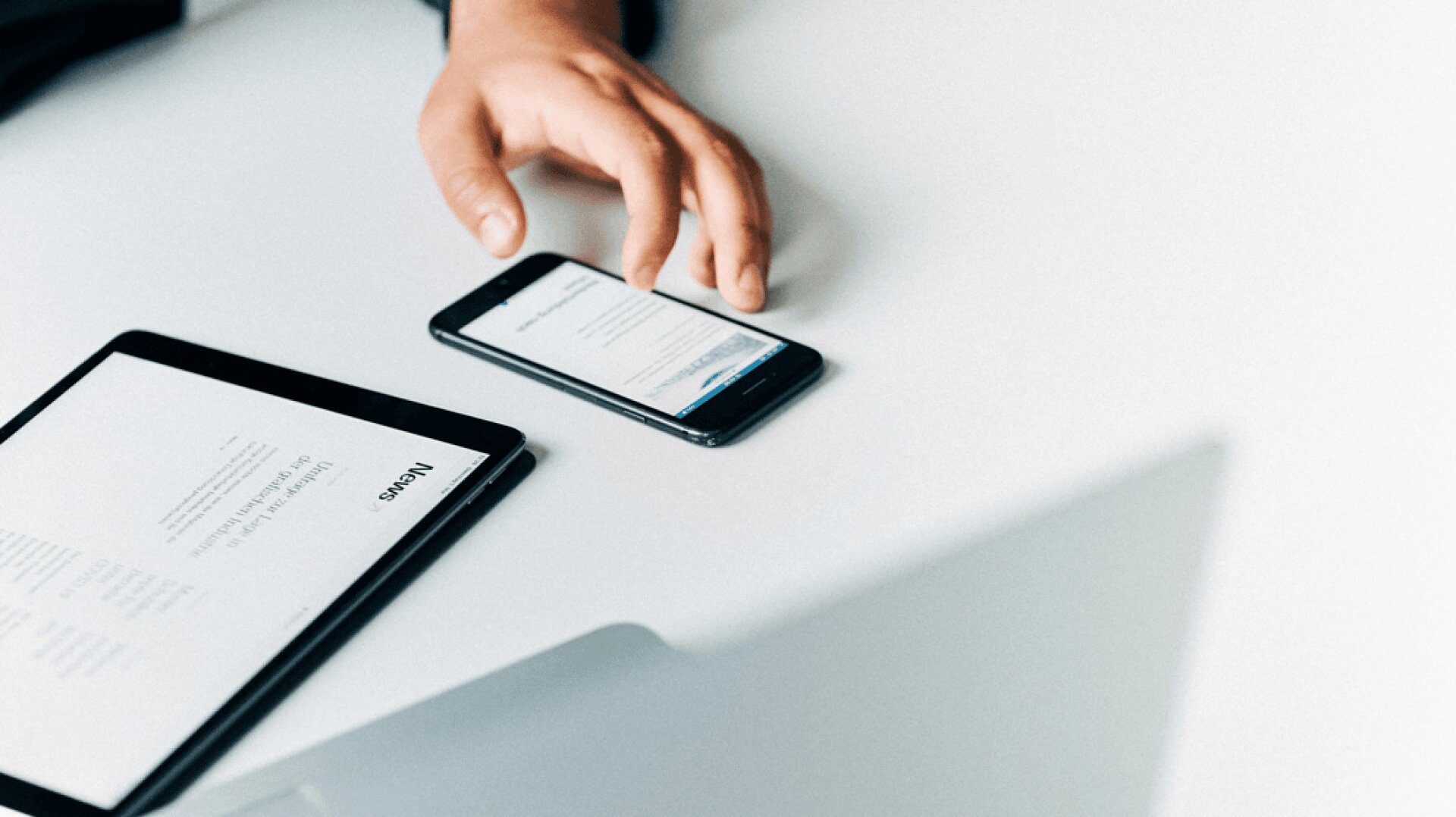 Die Hand einer Person ruht auf einem Smartphone, das Text anzeigt, neben einem Tablet mit einem geöffneten Dokument auf einem weißen Schreibtisch. Im Vordergrund ist ein verschwommener Laptop zu sehen.