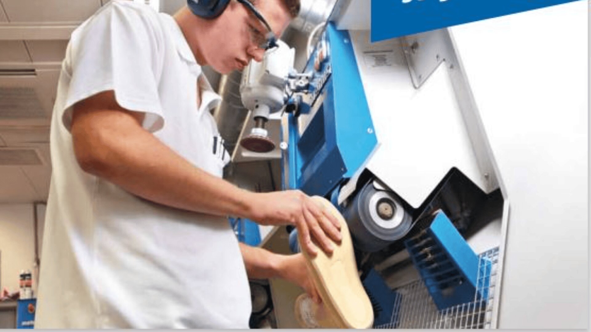 Eine Person, die eine Schutzbrille und einen Gehörschutz trägt, bedient eine Maschine und formt oder poliert eine Schuhsohle in einer scheinbaren Produktions- oder Werkstattumgebung.