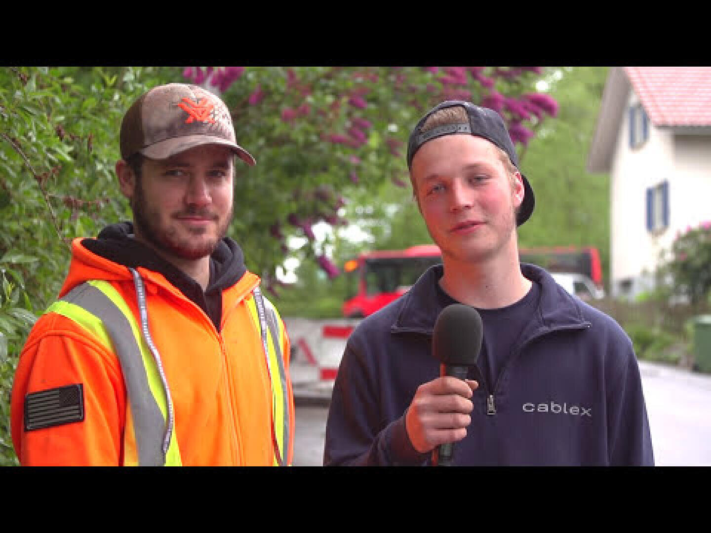 Zwei junge Männer stehen im Freien; der eine trägt eine orangefarbene Sicherheitsjacke und Mütze, der andere eine marineblaue Jacke und eine umgekehrte Mütze und hält ein Mikrofon in der Hand. Im Hintergrund sind grünes Laub und ein rotes Fahrzeug zu sehen.