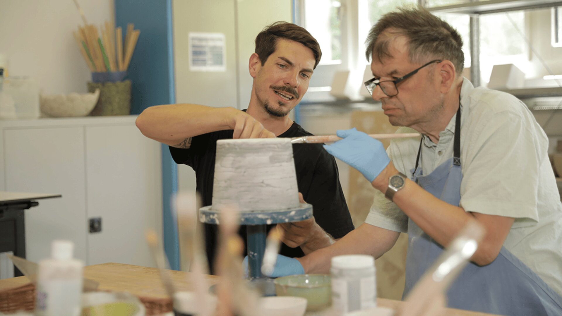 Zwei Männer arbeiten zusammen in einer Töpferwerkstatt, wobei der eine den anderen beim Bemalen eines Tontopfes anleitet. Der Ausbilder lächelt, während der andere blaue Handschuhe und eine Schürze trägt und sich auf das Bemalen des Topfes konzentriert. Auf dem Tisch liegen Malutensilien.