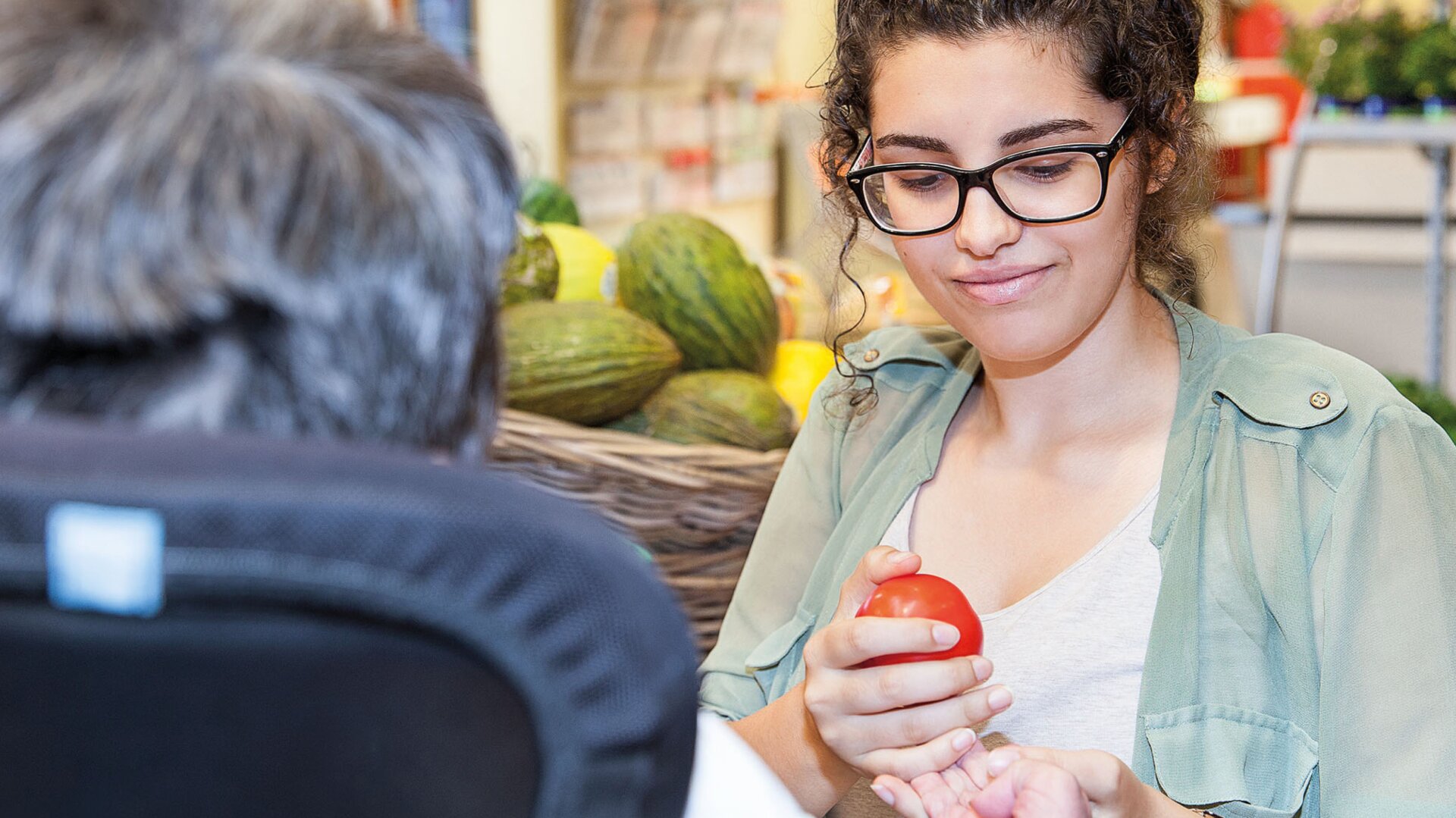 Eine junge Frau mit Brille reicht einer älteren Person im Rollstuhl beim Einkaufen eine Tomate, während im Hintergrund Körbe mit Lebensmitteln zu sehen sind.