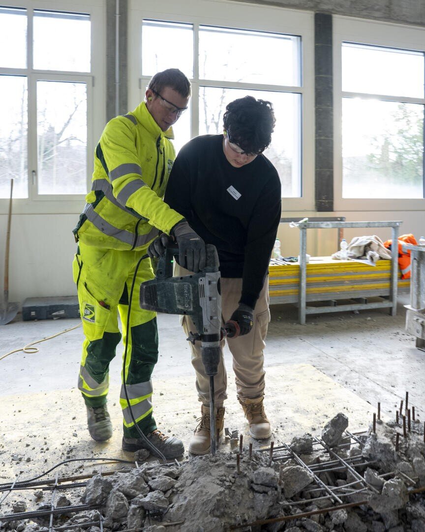 Zwei Personen arbeiten in einem Haus mit einem Presslufthammer und brechen Beton auf. Der eine trägt einen gelben Sicherheitsanzug und führt den anderen, der in schwarz-hellbrauner Kleidung gekleidet ist. Im Hintergrund sind Baumaterialien zu sehen.