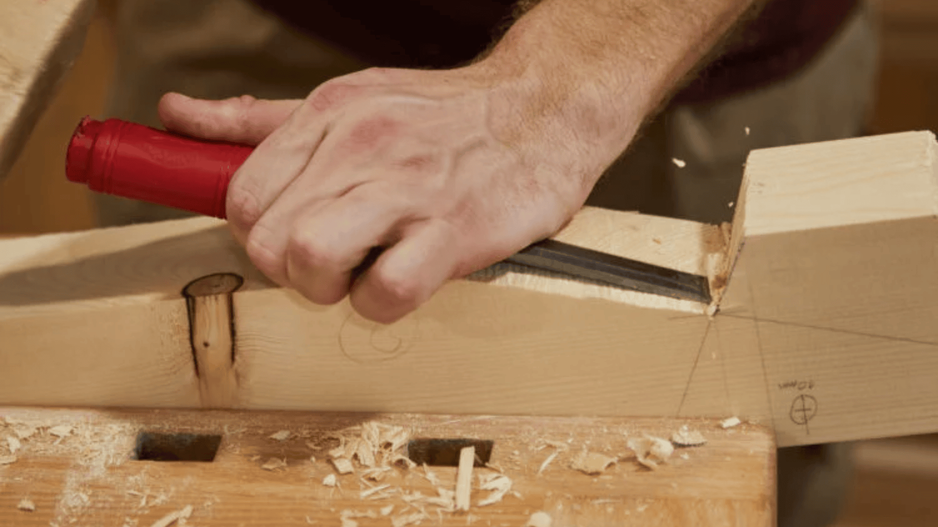 Eine Person schnitzt mit einem Meißel mit rotem Griff eine Kerbe in ein Stück helles Holz, das auf einer Werkbank liegt, wobei Holzspäne verstreut werden.