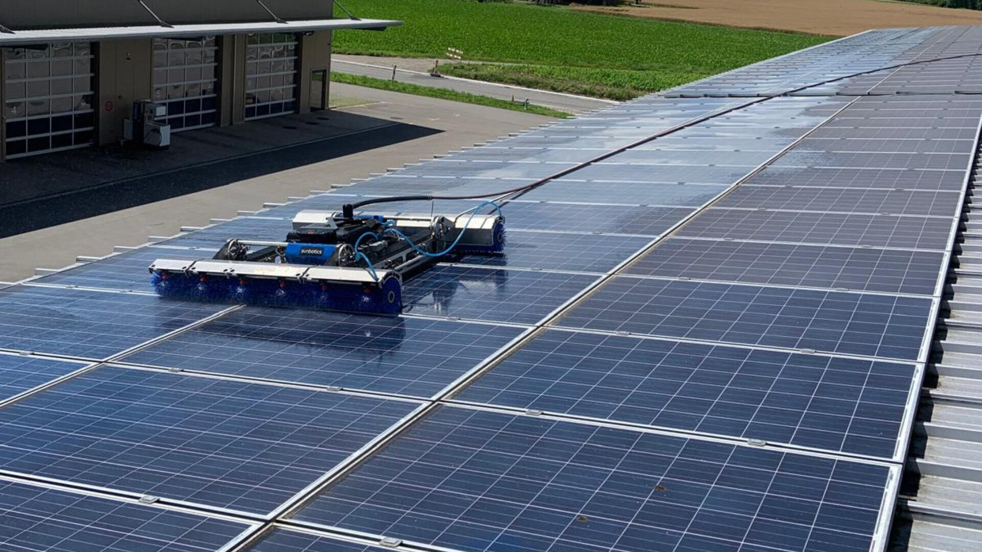 Ein Reinigungsroboter bewegt sich über Reihen von Solarmodulen auf einem großen Dach und entfernt Schmutz, um die Effizienz zu erhalten. Im Hintergrund sind ein Gebäude und grüne Felder zu sehen.