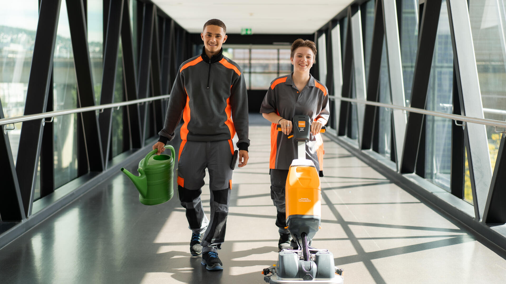 Zwei lächelnde Arbeiter in grauen und orangefarbenen Uniformen gehen einen gläsernen Gang entlang; einer hält eine grüne Gießkanne, der andere bedient eine Bodenreinigungsmaschine.