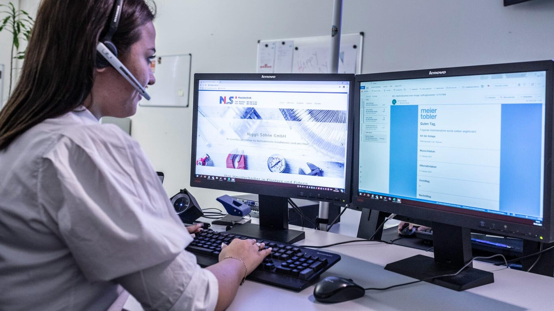 Eine Frau mit einem Headset arbeitet an einem Schreibtisch mit zwei Computermonitoren, von denen einer eine Website und der andere eine E-Mail anzeigt. Sie tippt auf einer Tastatur in einer Büroumgebung.