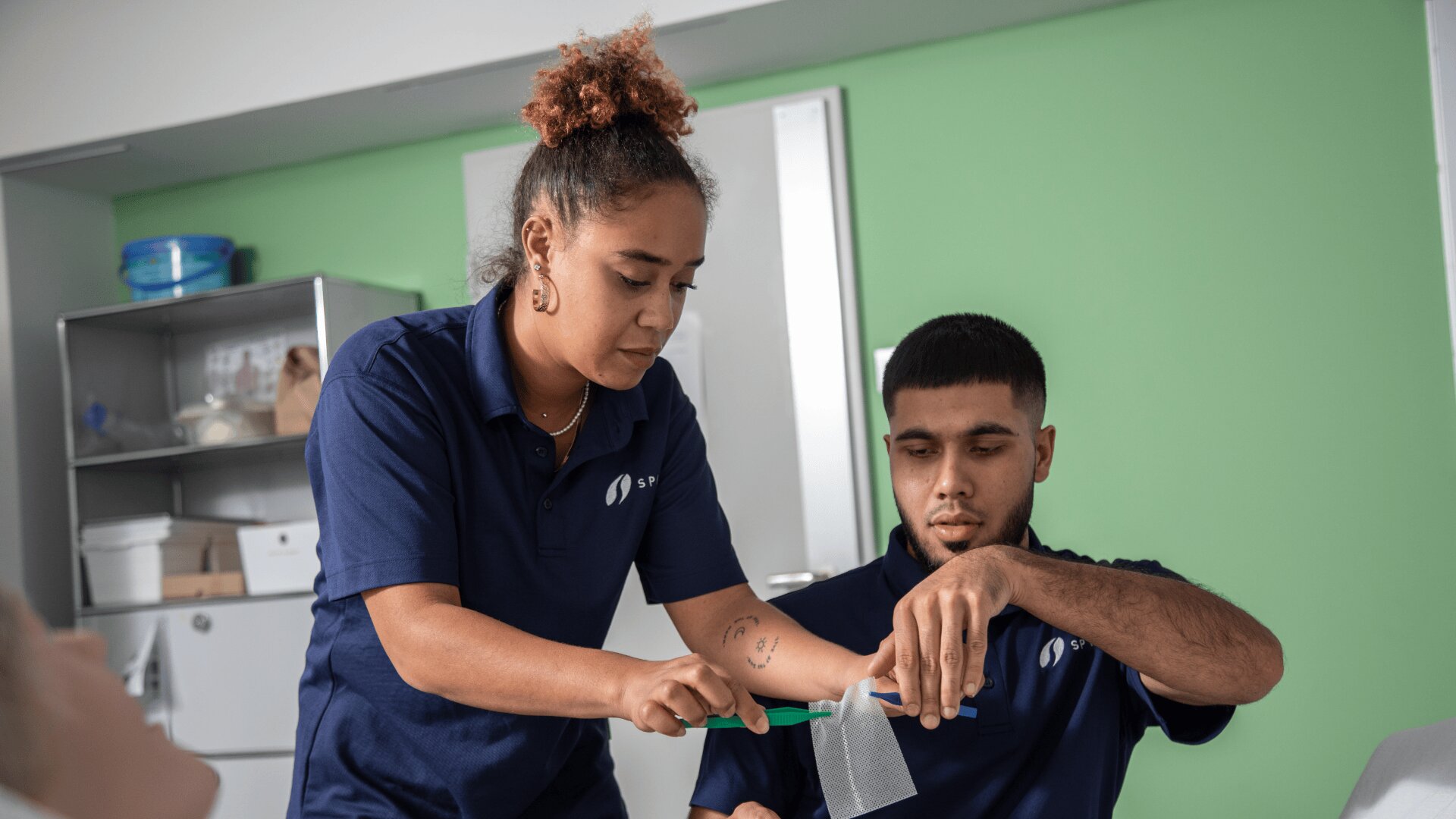 Eine Frau in einem marineblauen Polohemd wickelt einem Mann einen Verband um die Hand, während er sie festhält. Sie befinden sich in einem Innenraum, in einem medizinischen oder schulischen Umfeld mit grünen und weißen Wänden.