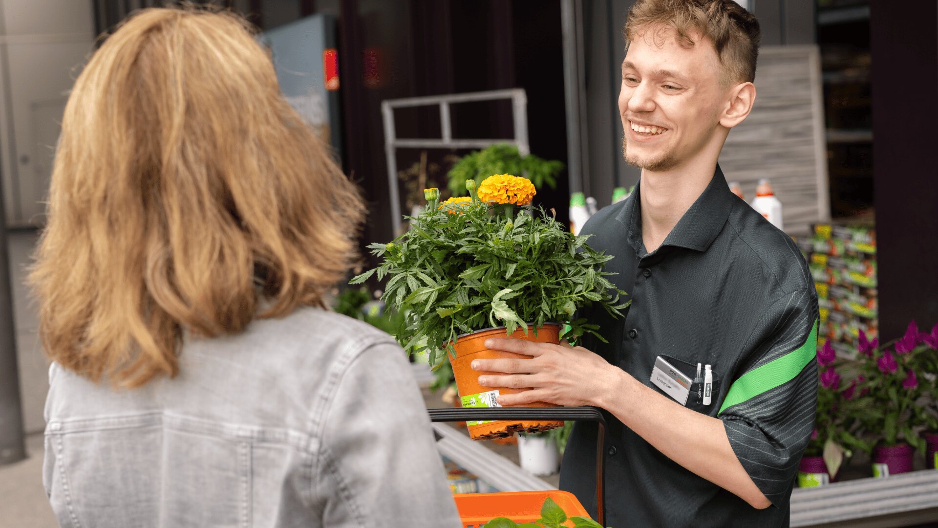Ein lächelnder Angestellter eines Gartencenters überreicht einer Kundin in einer grauen Jacke eine getopfte Ringelblumenpflanze.