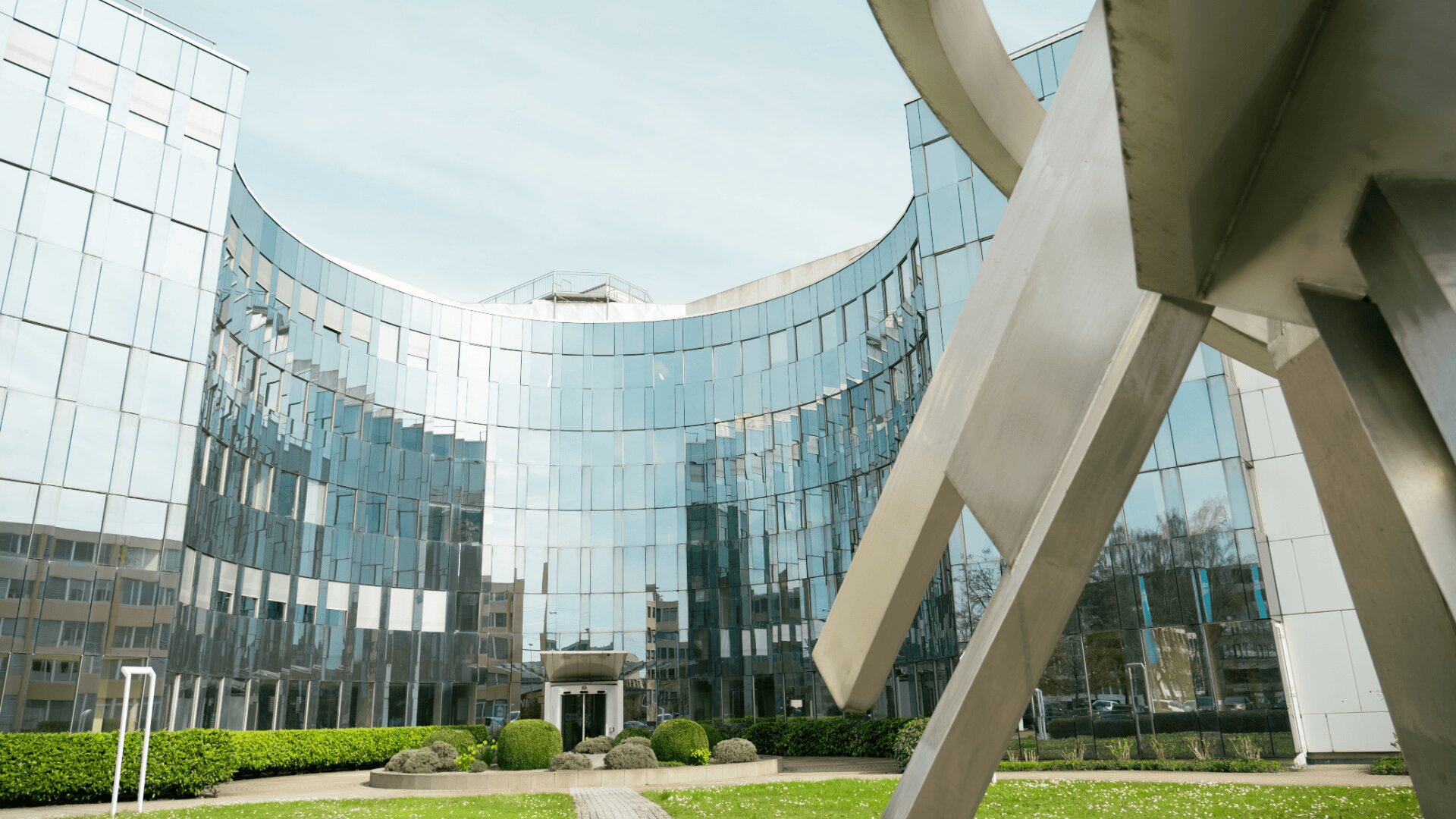 Gezeigt wird ein modernes gläsernes Bürogebäude mit einer geschwungenen Fassade, in der sich der Himmel und die umliegenden Gebäude spiegeln. Im Vordergrund befindet sich eine große abstrakte Metallskulptur auf einer grünen Rasenfläche.