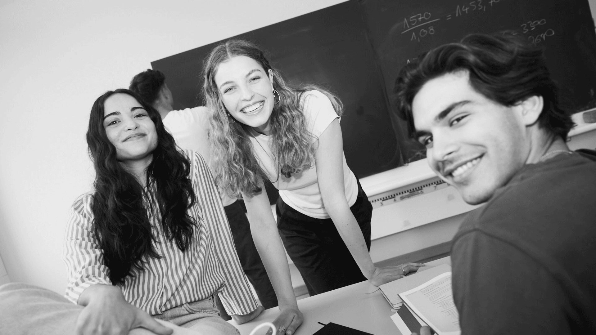 Drei junge Erwachsene lächeln und posieren gemeinsam in einem Klassenzimmer mit einer Kreidetafel und offenen Büchern auf dem Schreibtisch hinter ihnen. Die Atmosphäre scheint freundlich und kooperativ zu sein.