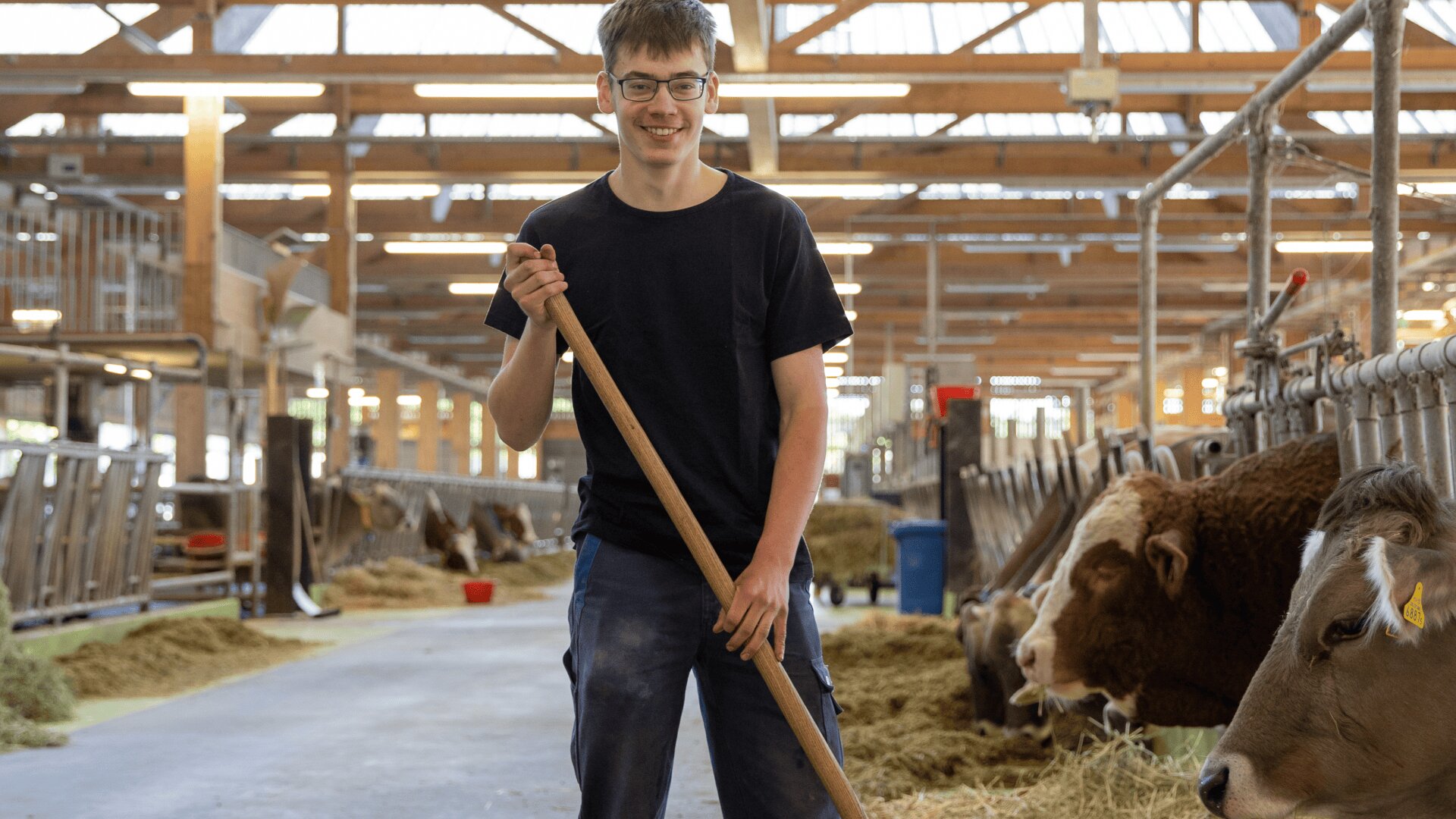 Ein junger Mann mit Brille und schwarzem Hemd steht lächelnd in einer Scheune und hält einen Besen in der Hand. In den Ställen neben ihm fressen die Kühe Heu. Die Scheune hat eine hohe Holzdecke und ist gut beleuchtet.