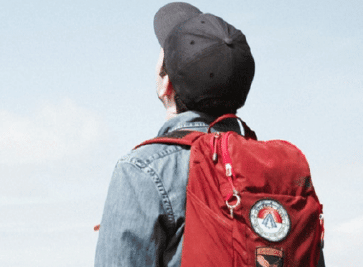 Eine Person, die eine dunkle Mütze, eine Jeansjacke und einen roten Rucksack trägt, blickt in den Himmel. Der Rucksack hat bunte Aufnäher. Der Hintergrund ist meist klarer Himmel.