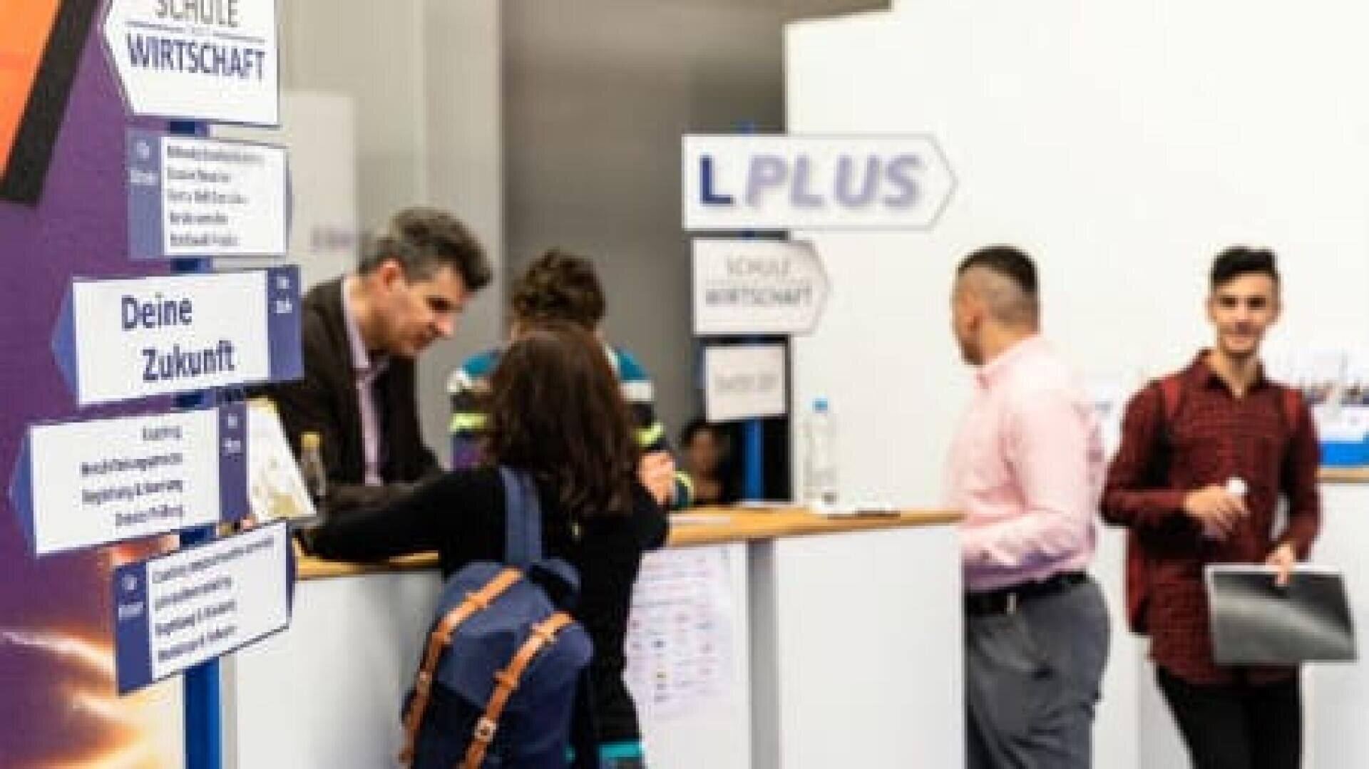 Mehrere Personen unterhalten sich an einem Informationsstand mit der Aufschrift Schule Wirtschaft und L Plus. Schilder und Flugblätter sind zu sehen, und eine Person im Vordergrund geht lächelnd vorbei, mit einem Rucksack und Papieren.