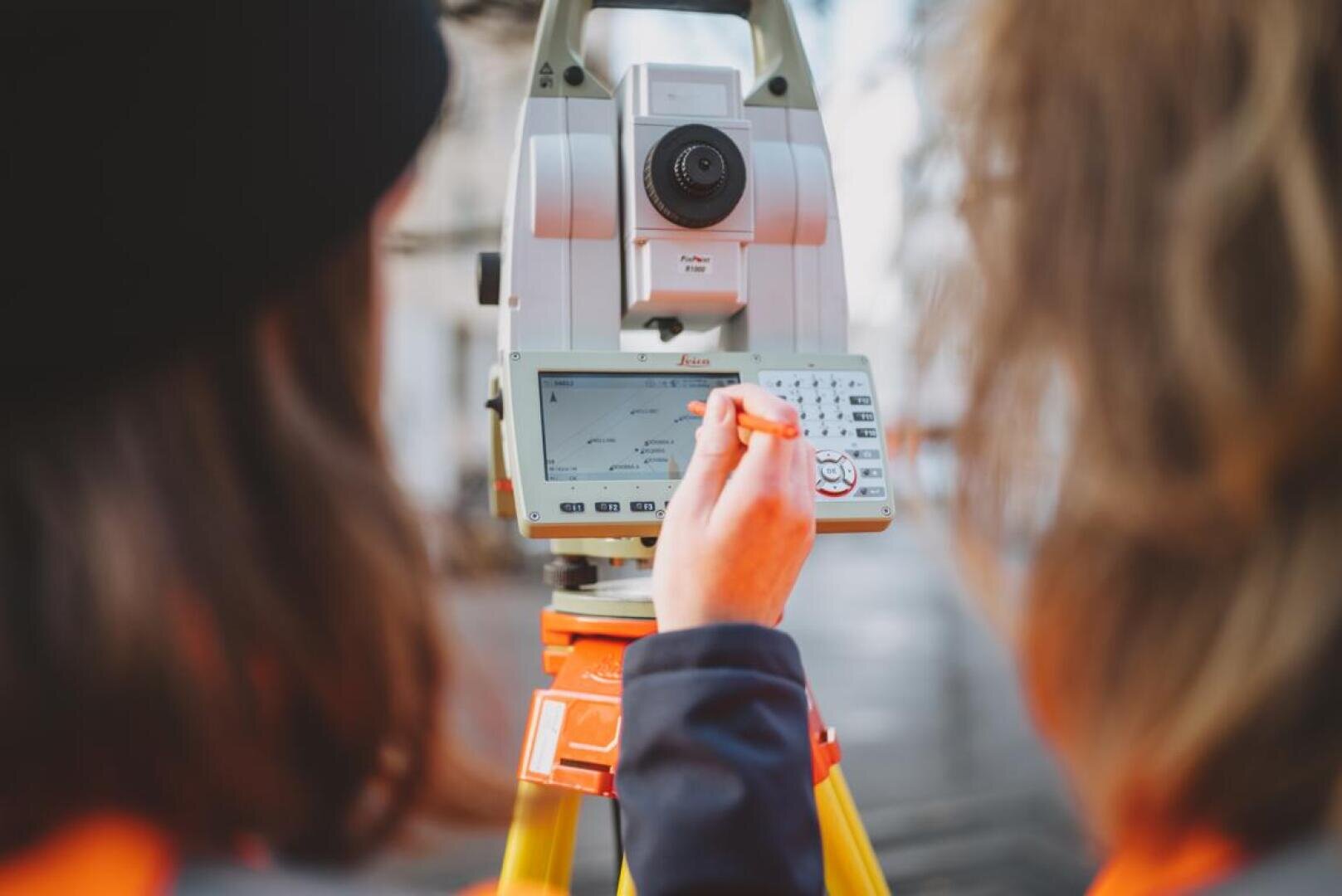 Zwei Personen in orangefarbener Kleidung bedienen ein Vermessungsinstrument, wobei eine Person einen Stift auf dem Touchscreen verwendet. Der Fokus liegt auf dem Display und den Bedienelementen des Geräts, wobei die beiden Personen im Vordergrund leicht unscharf sind.