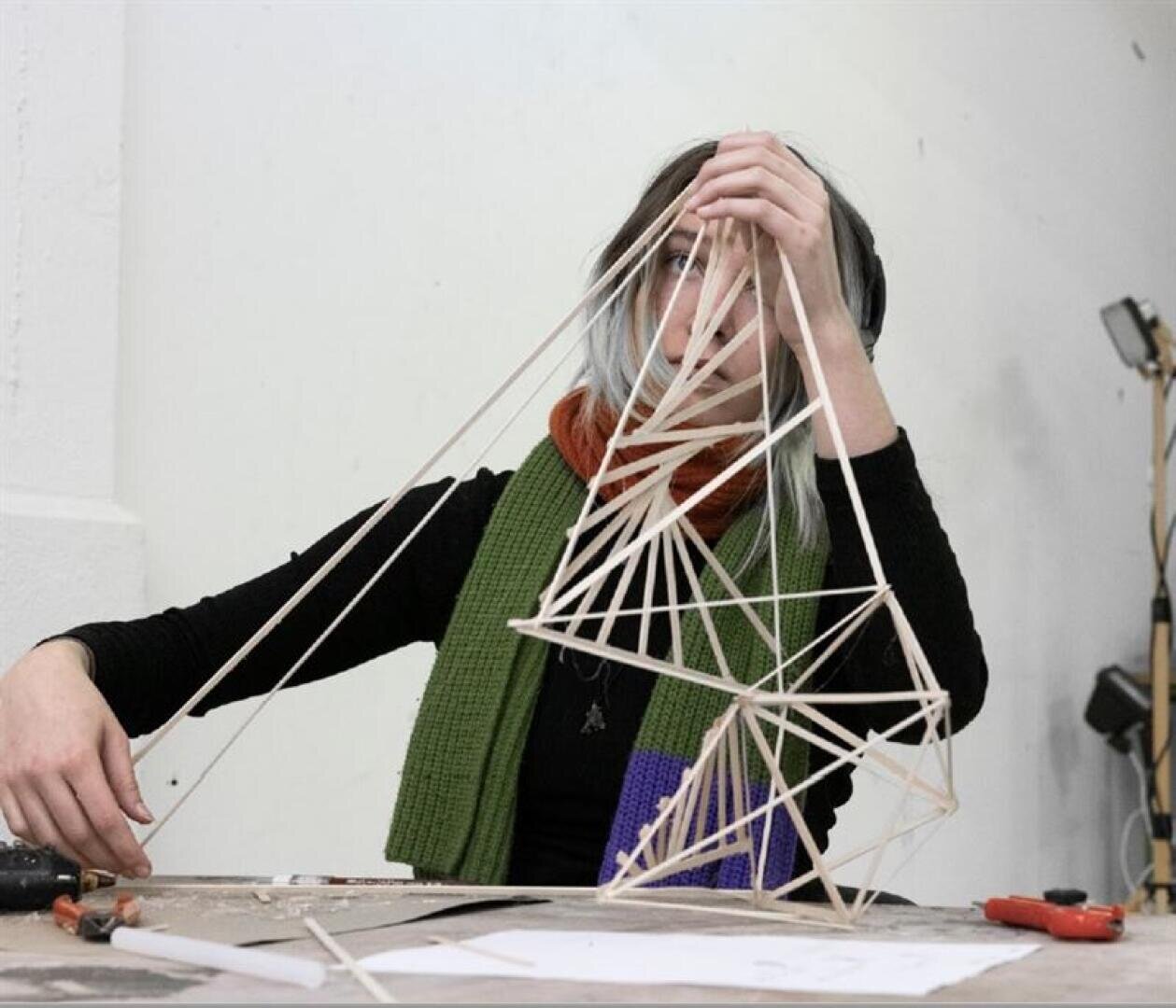 Eine Person mit schulterlangem Haar hält ein geometrisches Gebilde aus Holzstäben und Schnüren hoch, während sie an einem Tisch mit verstreuten Papieren und Werkzeugen sitzt.