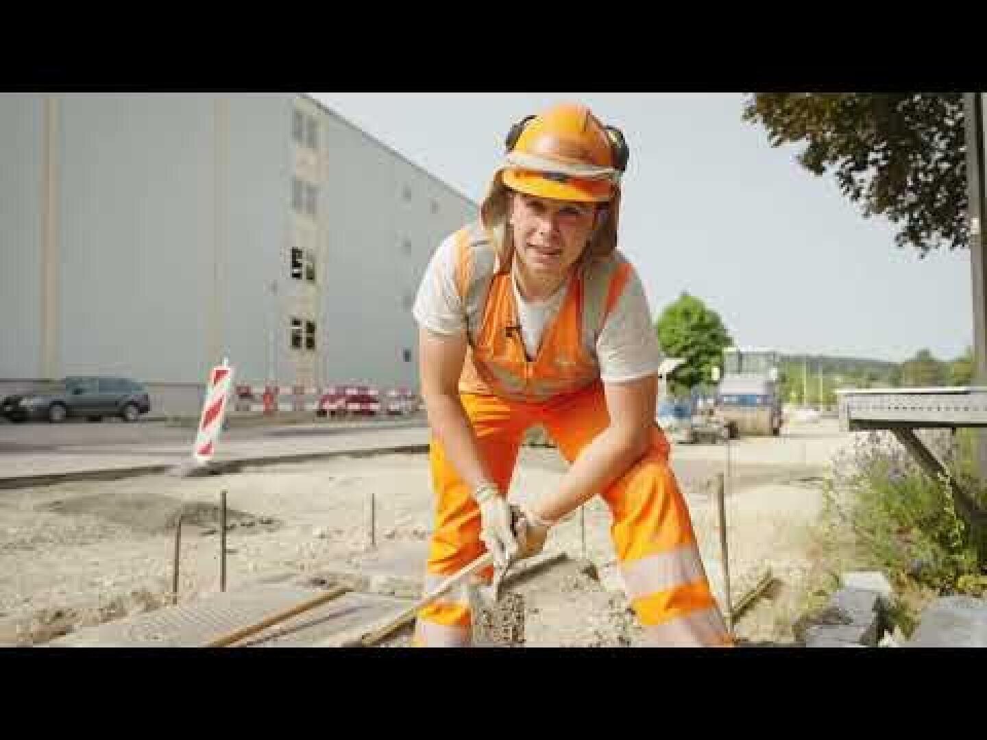 Ein Bauarbeiter in orangefarbener Schutzkleidung und mit Helm bückt sich und hält ein Metallwerkzeug auf einer Baustelle; im Hintergrund sind ein Gebäude und Fahrzeuge zu sehen.