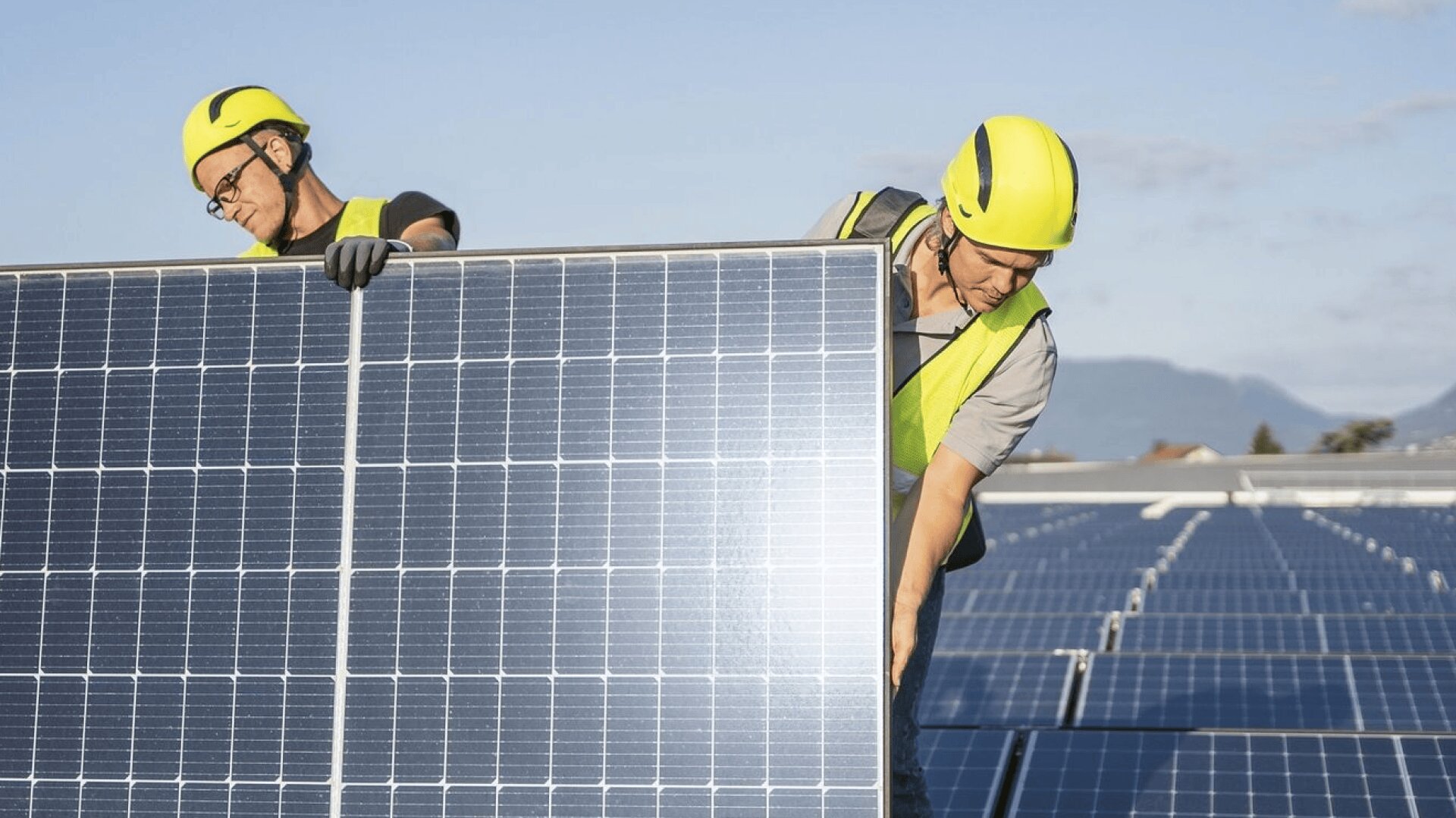 Zwei Arbeiter mit gelben Helmen und Sicherheitswesten installieren ein großes Solarmodul auf einem Dach, während im Hintergrund bei klarem Himmel Reihen von Solarmodulen zu sehen sind.