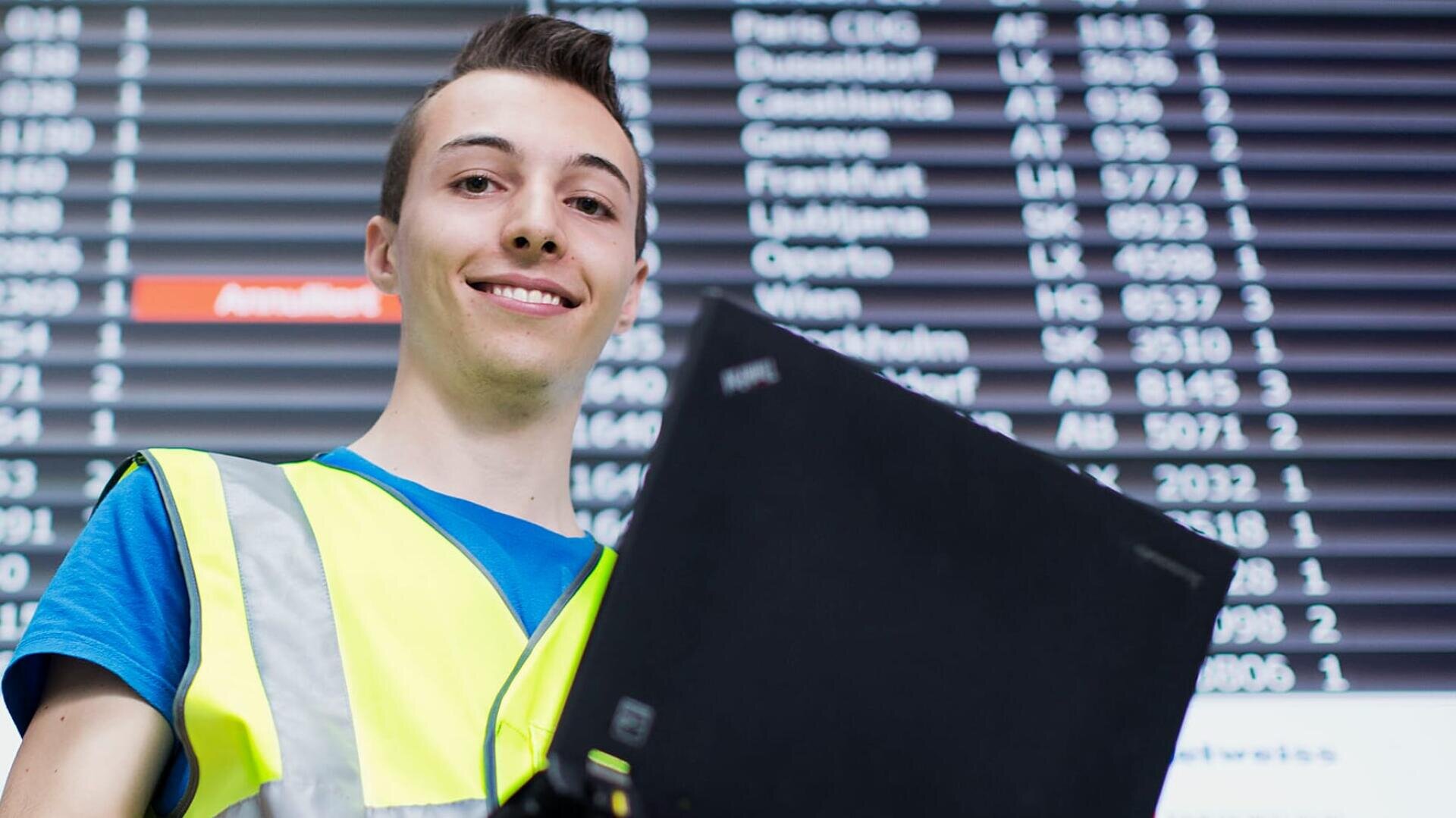 Eine lächelnde Person in einer gelben Sicherheitsweste hält einen Laptop vor einer großen digitalen Flughafen-Abflugtafel, auf der Fluginformationen angezeigt werden.