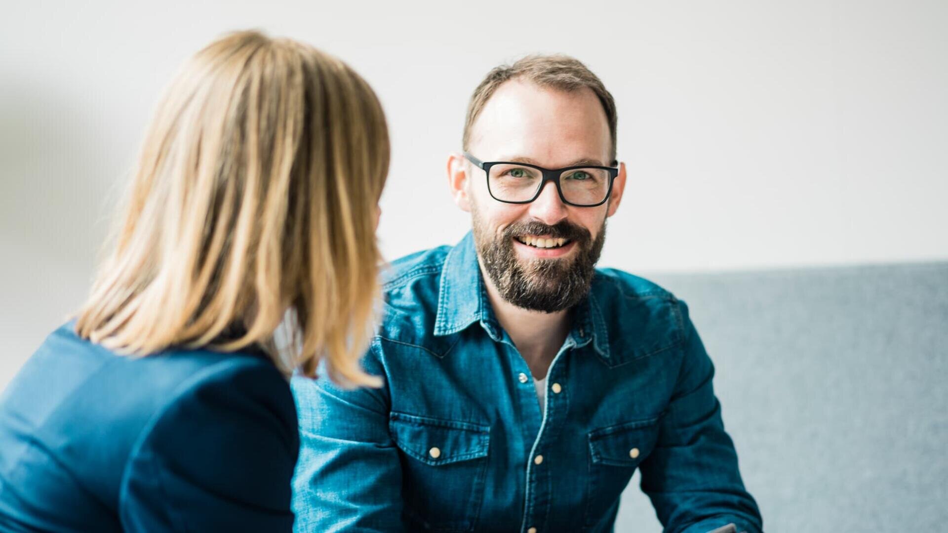Ein bärtiger Mann mit Brille und Jeanshemd lächelt, während er sich mit einer Frau mit blondem Haar und blauem Blazer unterhält. Beide sitzen in einem Innenraum auf einem grauen Sofa.