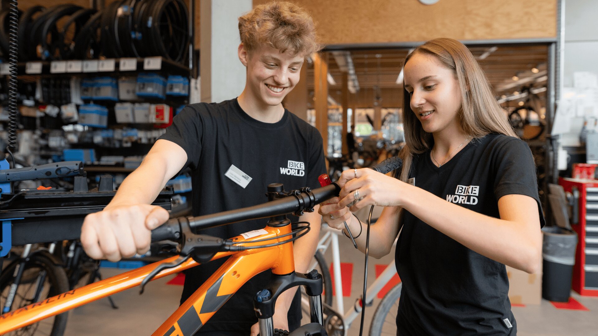 Zwei junge Leute in "Bike World"-Shirts reparieren gemeinsam den Lenker eines orangefarbenen Fahrrads in einem Fahrradgeschäft. Im Hintergrund sind Regale mit Werkzeug und Fahrradteilen zu sehen.