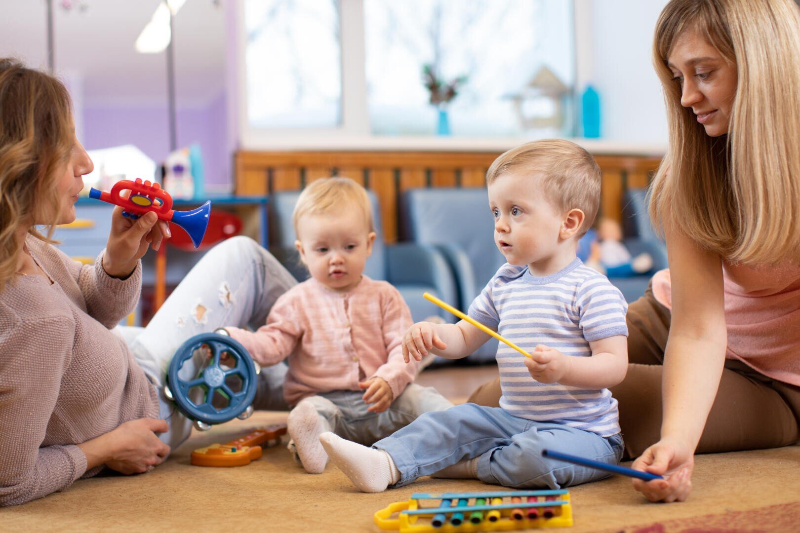 Zwei Frauen und zwei Kleinkinder sitzen in einem hellen, gemütlichen Raum mit großen Fenstern im Hintergrund auf dem Boden und spielen mit Musikspielzeug, darunter eine Spielzeug-Trompete, eine Trommel und ein Xylophon.