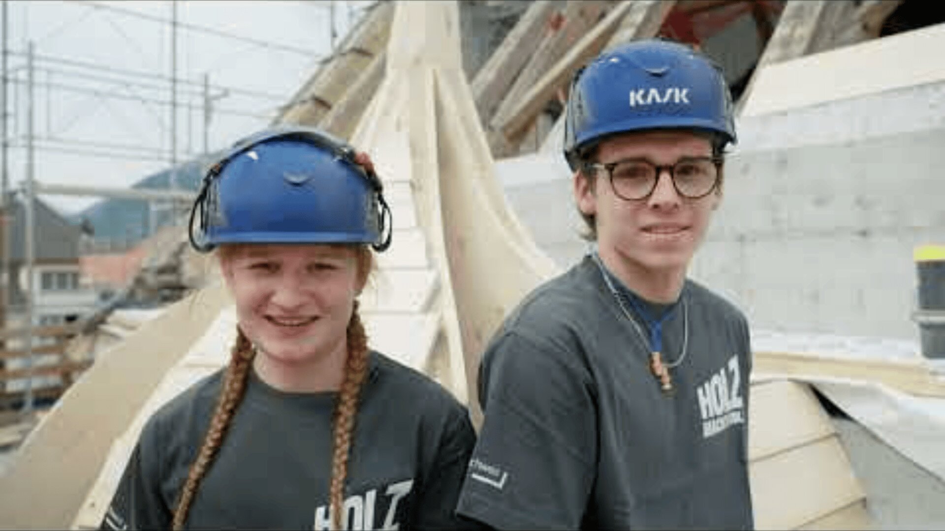 Zwei junge Leute mit blauen Schutzhelmen und grauen HOLZ-Hemden stehen lächelnd nebeneinander auf einer Baustelle, auf der im Hintergrund Holzbalken und Gerüste zu sehen sind.