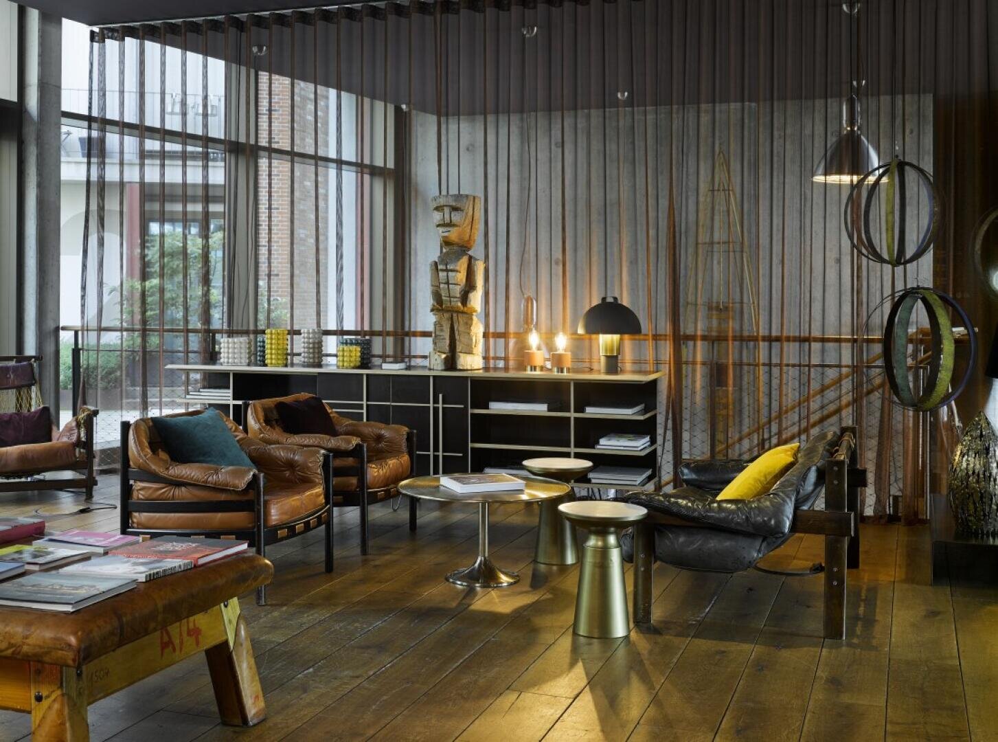 Ein modernes Wohnzimmer mit Ledersesseln, einem hölzernen Couchtisch, Skulpturen, raumhohen Fenstern und eklektischem Dekor. Warme Beleuchtung und verschiedene gelbe und grüne Akzente schaffen eine gemütliche, stilvolle Atmosphäre.