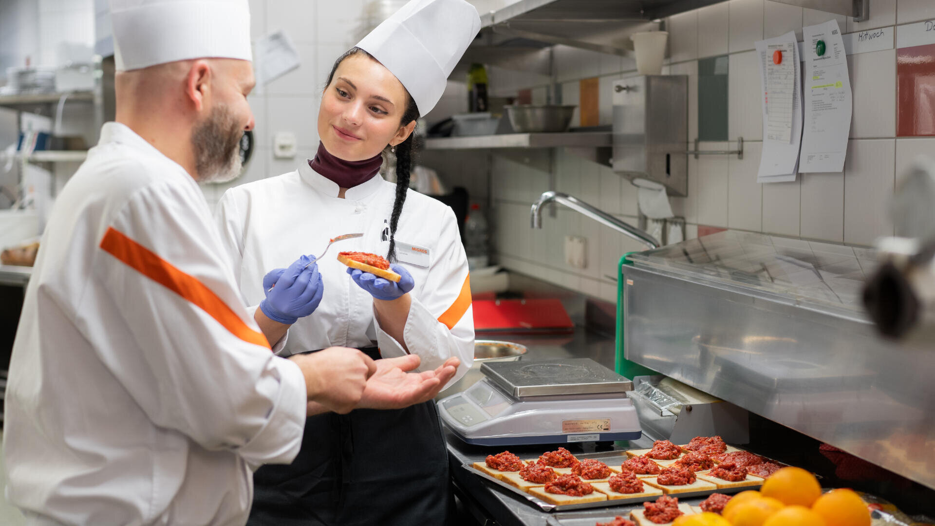 Zwei Köche in einer Großküche diskutieren über ein Stück Bruschetta. Die Frau hält die Schale und lächelt ihren Kollegen an. Auf der Theke neben ihnen sind mehrere Bruschetta-Stücke und Orangen zu sehen.
