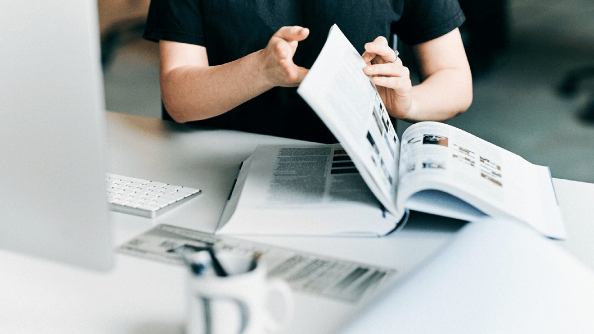 Eine Person in einem schwarzen Hemd sitzt an einem Schreibtisch und blättert in einem Buch mit Bildern und Text. Ein aufgeschlagenes Buch, eine Computertastatur und eine weiße Tasse stehen ebenfalls auf dem Schreibtisch. Die Szene scheint ein Arbeitszimmer oder eine Arbeitsumgebung zu sein.