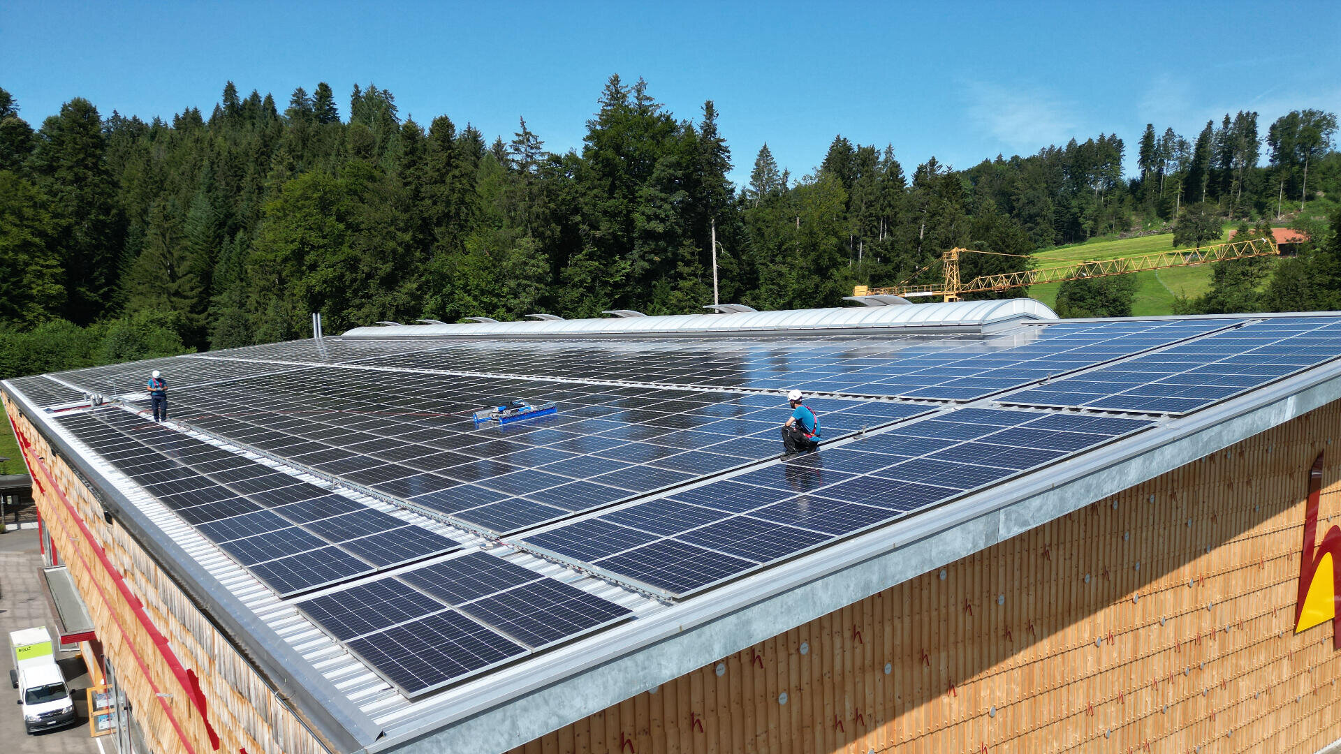 Arbeiter in Schutzkleidung reinigen und warten eine große Reihe von Solarmodulen auf einem hölzernen Gebäudedach, umgeben von grünen Bäumen und einem blauen Himmel im Hintergrund.