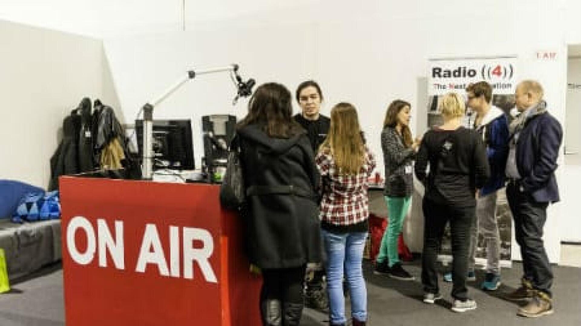 Eine Gruppe von Menschen steht und spricht in der Nähe eines Radiopults mit Mikrofonen und Computern. Auf dem roten Pult steht in großen weißen Buchstaben ON AIR. Im Hintergrund ist ein Radio (4)-Banner zu sehen.