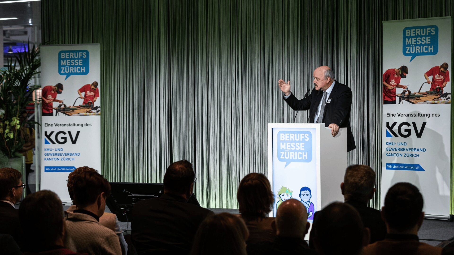 Ein Mann im Anzug spricht auf einem Podium auf einer Bühne während der Berufsmesse Zürich, im Vordergrund sind Banner und Publikum zu sehen.