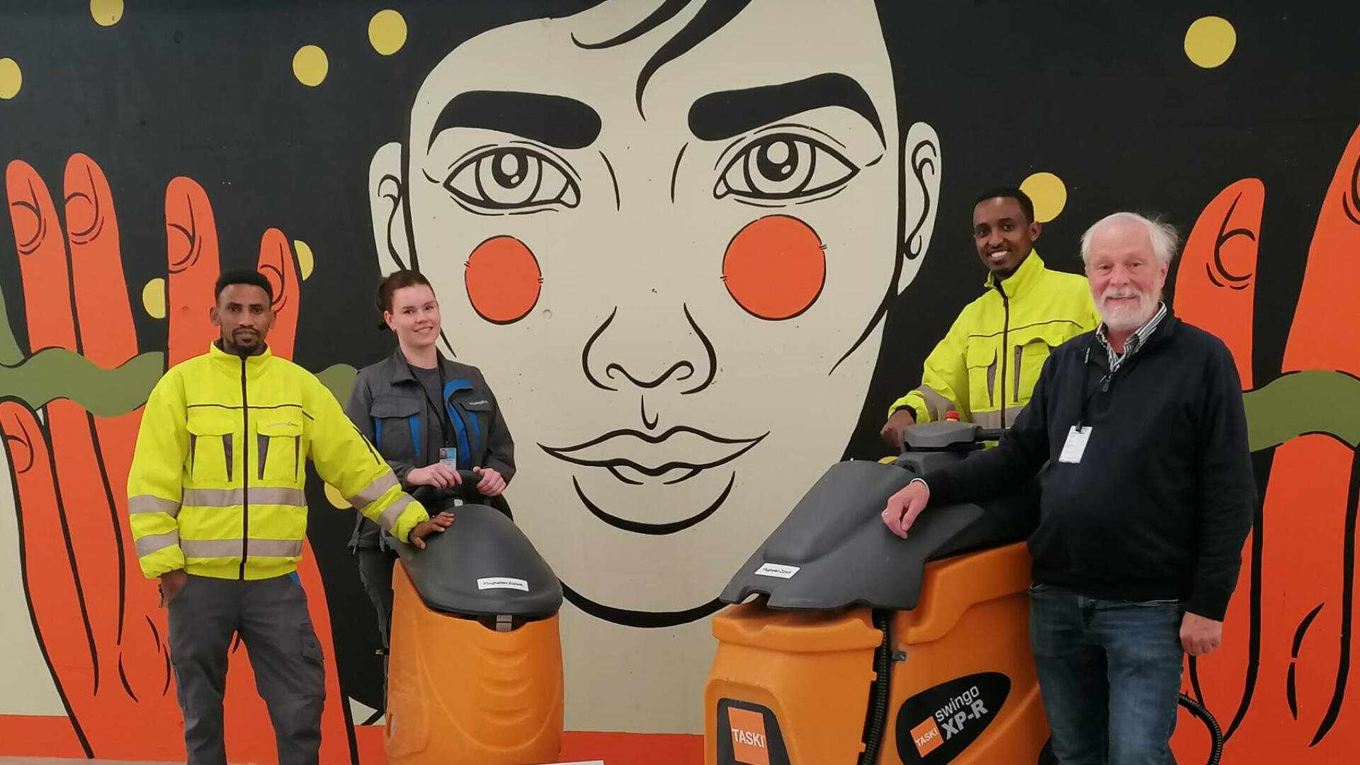 Vier Personen stehen vor einem bunten Wandgemälde mit einem großen Gesicht und Händen. Zwei der Personen tragen gelbe Jacken und zwei stehen an orangefarbenen industriellen Reinigungsmaschinen. Alle lächeln in die Kamera.