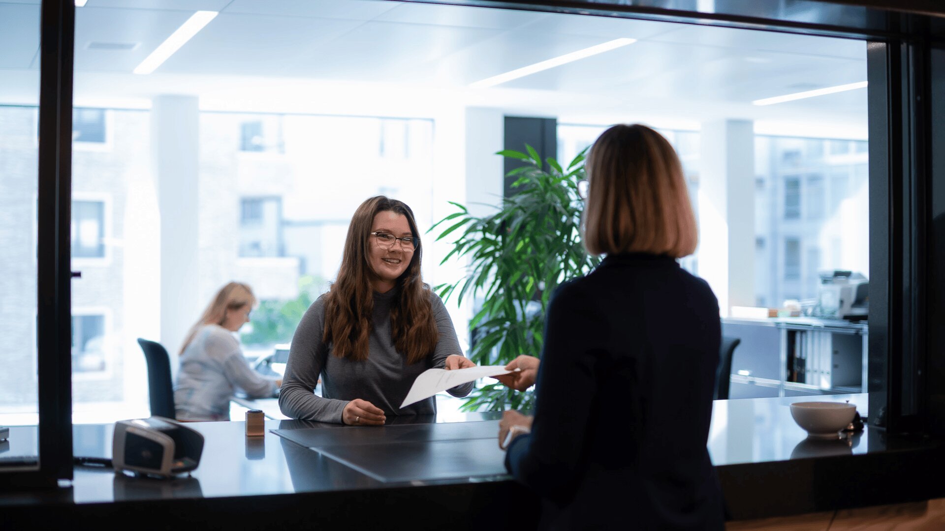 Eine Frau am Empfang überreicht einer anderen Frau, die ihr gegenüber steht, ein Dokument. Im Hintergrund arbeitet eine weitere Person an einem Computer in einem modernen, hellen Büro mit Pflanzen und großen Fenstern.