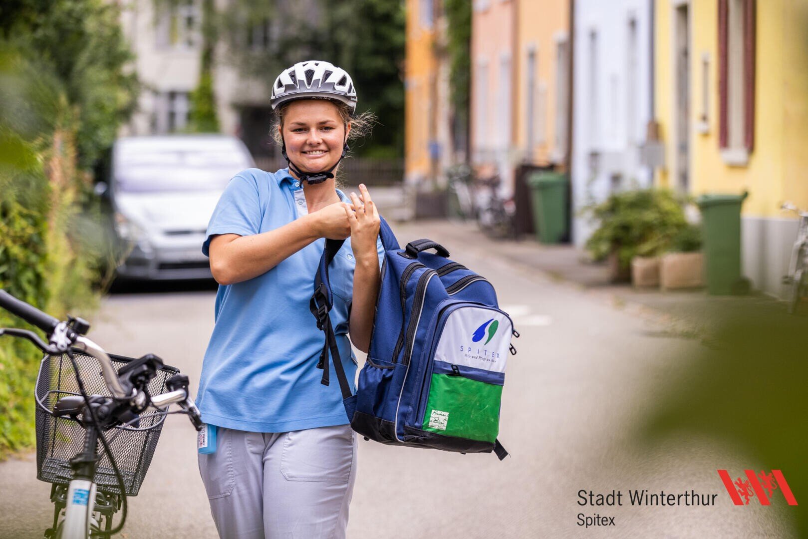 Eine lächelnde Frau in blauer Uniform und mit Helm fährt mit dem Velo durch eine Wohnstrasse und trägt einen Spitex-Rucksack. In der rechten unteren Ecke ist das Spitex-Logo der Stadt Winterthur zu sehen.