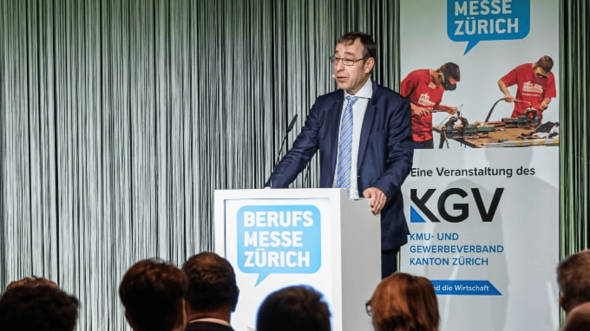 Ein Mann im Anzug spricht auf einem Podium mit der Aufschrift Berufsmesse Zürich vor einem Publikum. Im Hintergrund ist ein Banner zu sehen, auf dem Menschen arbeiten und Veranstaltungshinweise für die Berufsmesse Zürich zu sehen sind.