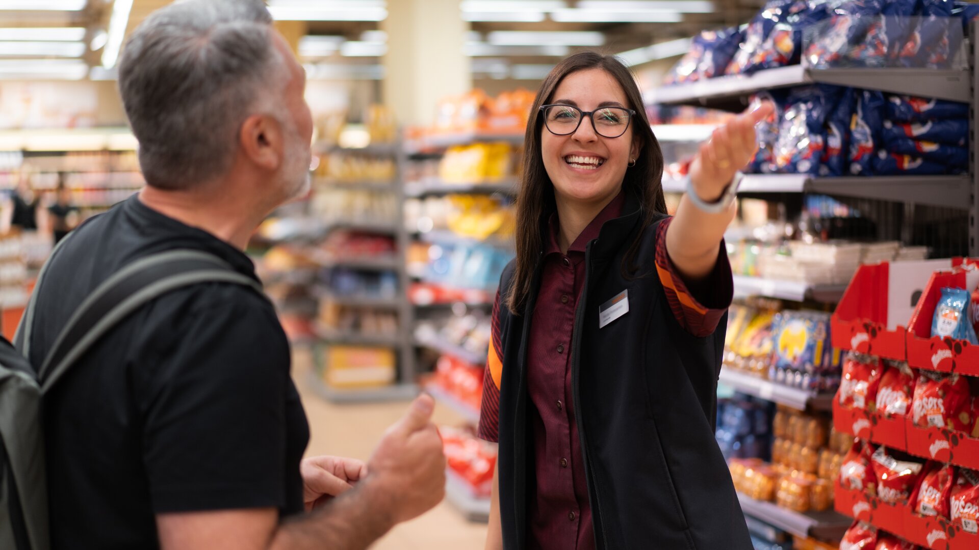 Eine lächelnde Verkäuferin hilft einem männlichen Kunden in einem mit Snacks und bunten Produkten gefüllten Lebensmittelgang mit Gesten.