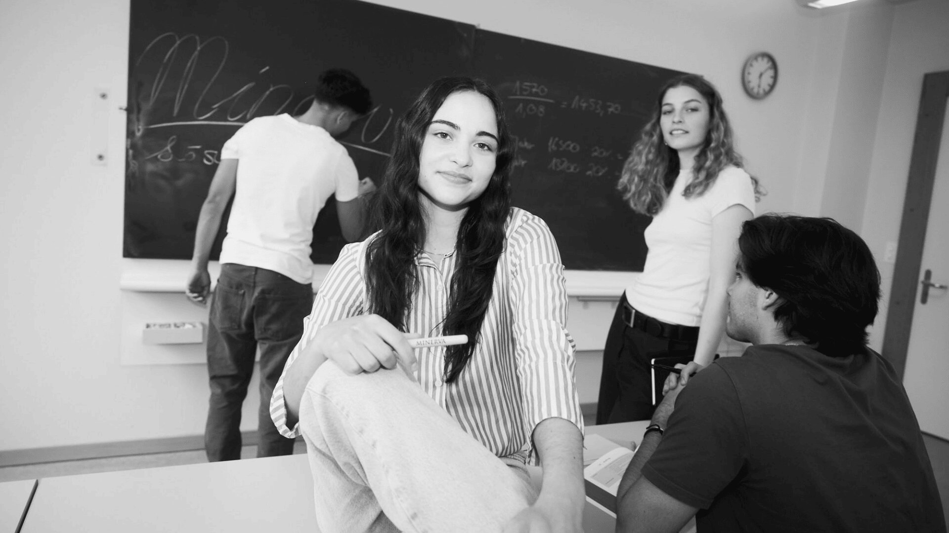 Eine Gruppe von vier Schülern in einem Klassenzimmer, von denen einer auf eine Kreidetafel schreibt, während die anderen sich unterhalten; eine junge Frau sitzt vorne und lächelt in die Kamera. Die Szene wirkt entspannt und gemeinschaftlich.
