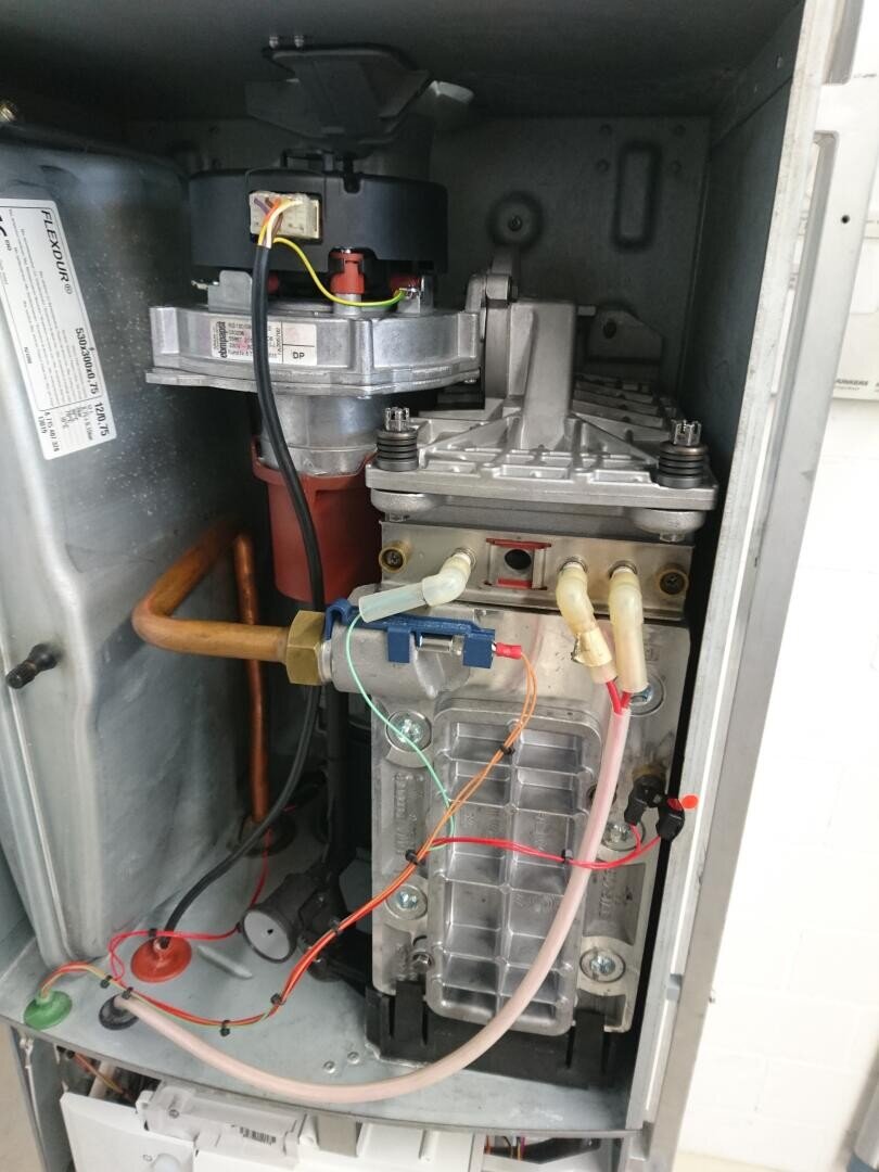 Das Bild zeigt das Innere eines Kessels oder einer Heizungsanlage mit sichtbaren Metallkomponenten, Kupferrohren, Drähten und Anschlüssen. Die mechanischen und elektrischen Innenteile sind zur Inspektion und Wartung zugänglich.