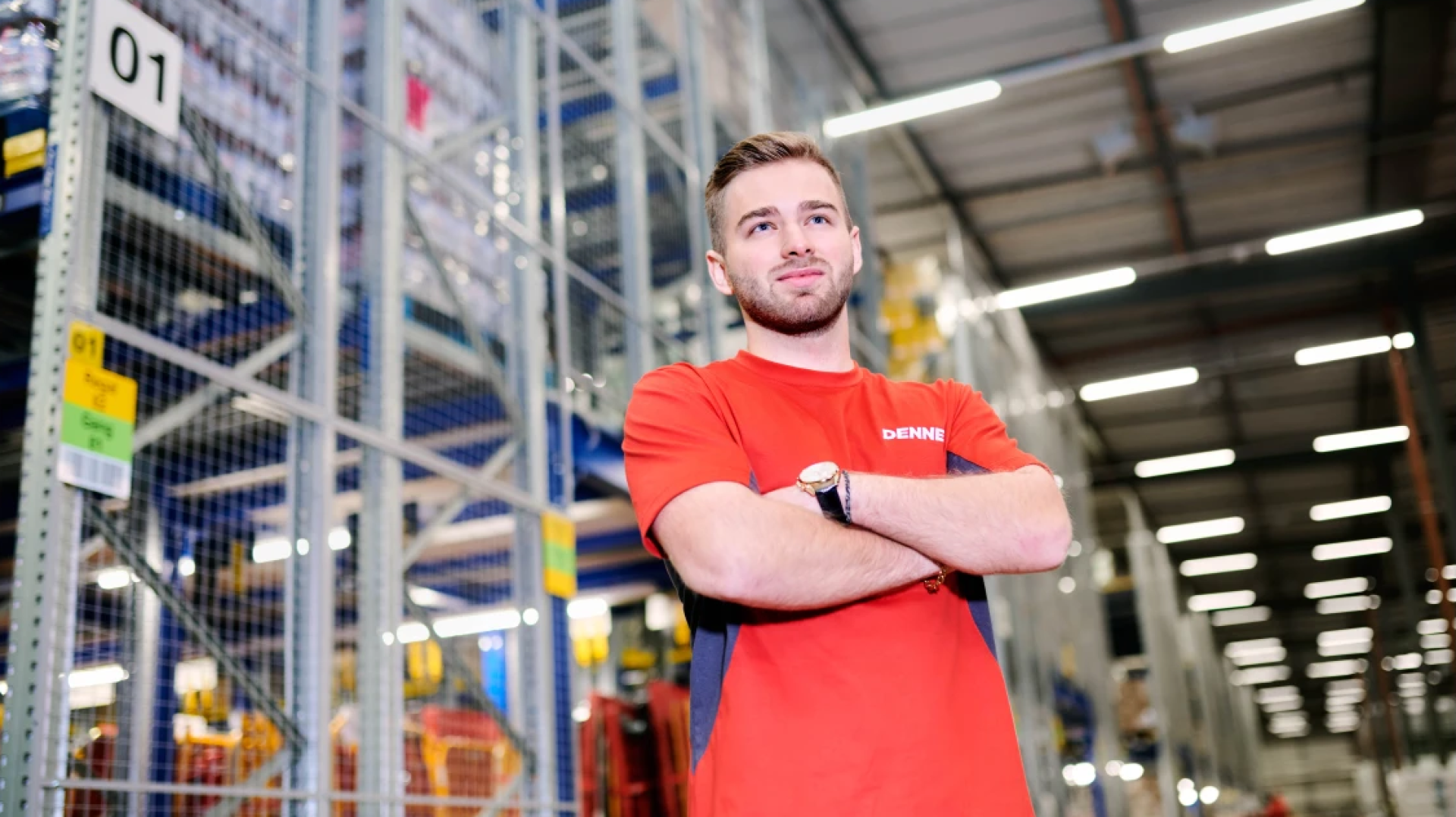 Ein Mann in einem roten Hemd steht selbstbewusst mit verschränkten Armen in einer großen Lagerhalle, umgeben von hohen Metallregalen, die mit Kisten gefüllt sind.