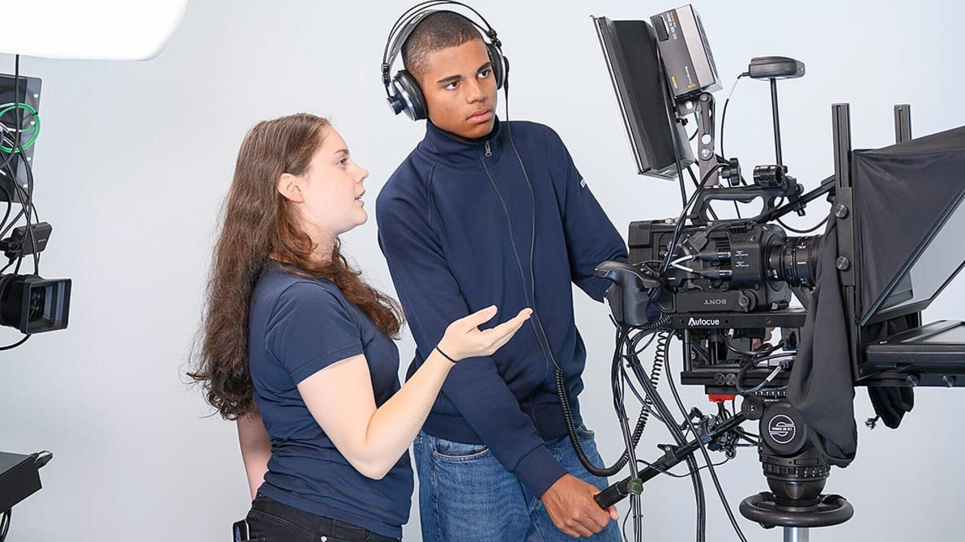 Zwei junge Menschen bedienen eine professionelle Videoausrüstung in einem Studio. Der eine trägt einen Kopfhörer und stellt die Regler ein, während der andere auf einen Monitor deutet. Beide sind auf den Drehvorgang konzentriert.