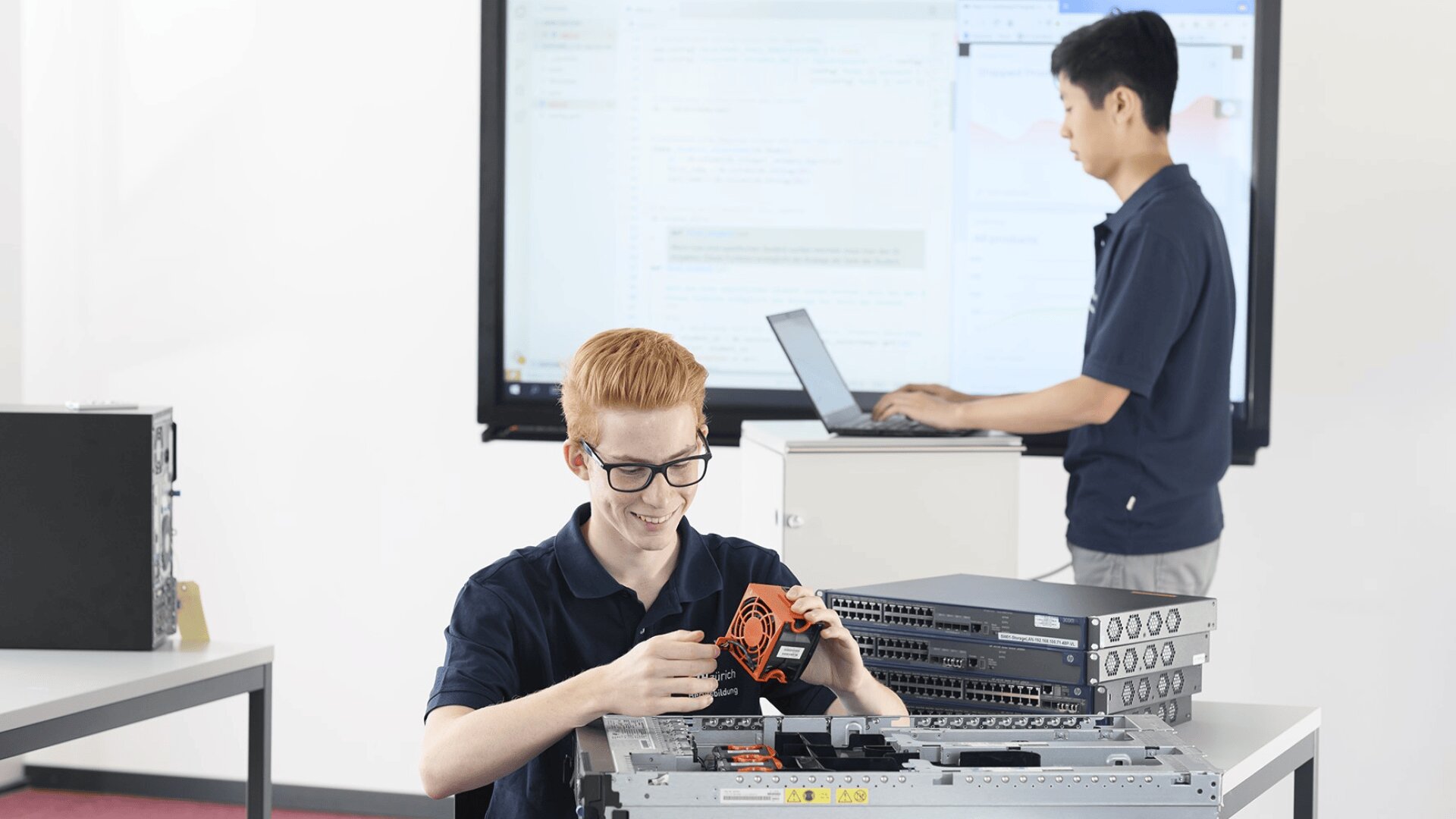 Ein junger Mann mit Brille arbeitet an einem Schreibtisch an Computerhardware, lächelt und hält ein Gerät. Eine andere Person steht im Hintergrund mit einem Laptop vor einem großen Bildschirm, auf dem ein Code angezeigt wird.
