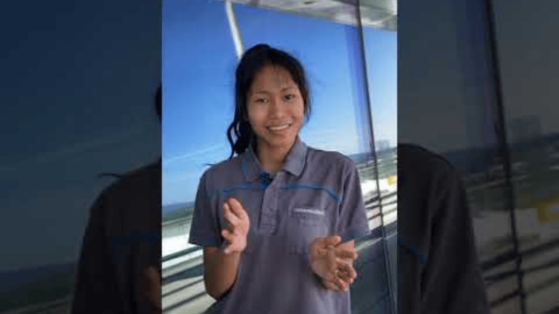 Eine Person mit langen dunklen Haaren und einem grauen Hemd mit Kragen steht draußen vor einer Glaswand, lächelt und gestikuliert mit den Händen. Im Hintergrund sind der blaue Himmel und das Geländer zu sehen.