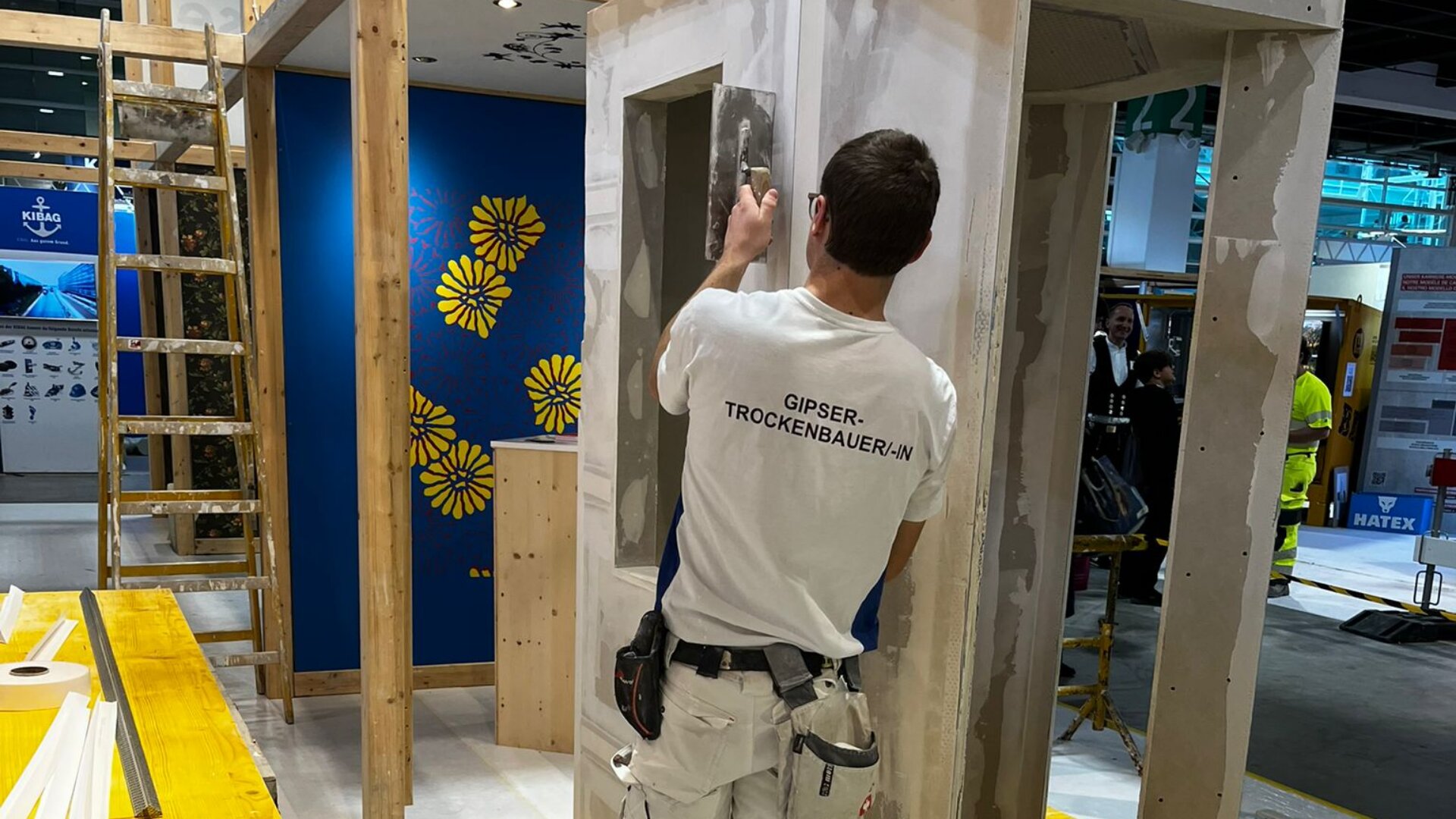 Ein Bauarbeiter in einem weißen Hemd schleift eine Trockenbauwand in einer Halle, umgeben von Holzbalken, Leitern und einer blauen Wand mit gelben Blumenmustern. An seinem Gürtel hängen Werkzeuge.