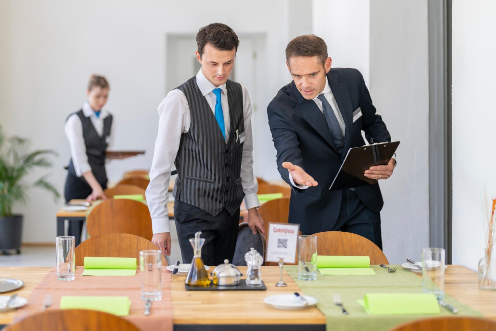 Zwei männliche Restaurantangestellte in Uniform stehen an einem gedeckten Tisch, wobei der eine ein Klemmbrett in der Hand hält und mit Gesten Anweisungen gibt. Im Hintergrund bedient eine Kellnerin an einem anderen Tisch.
