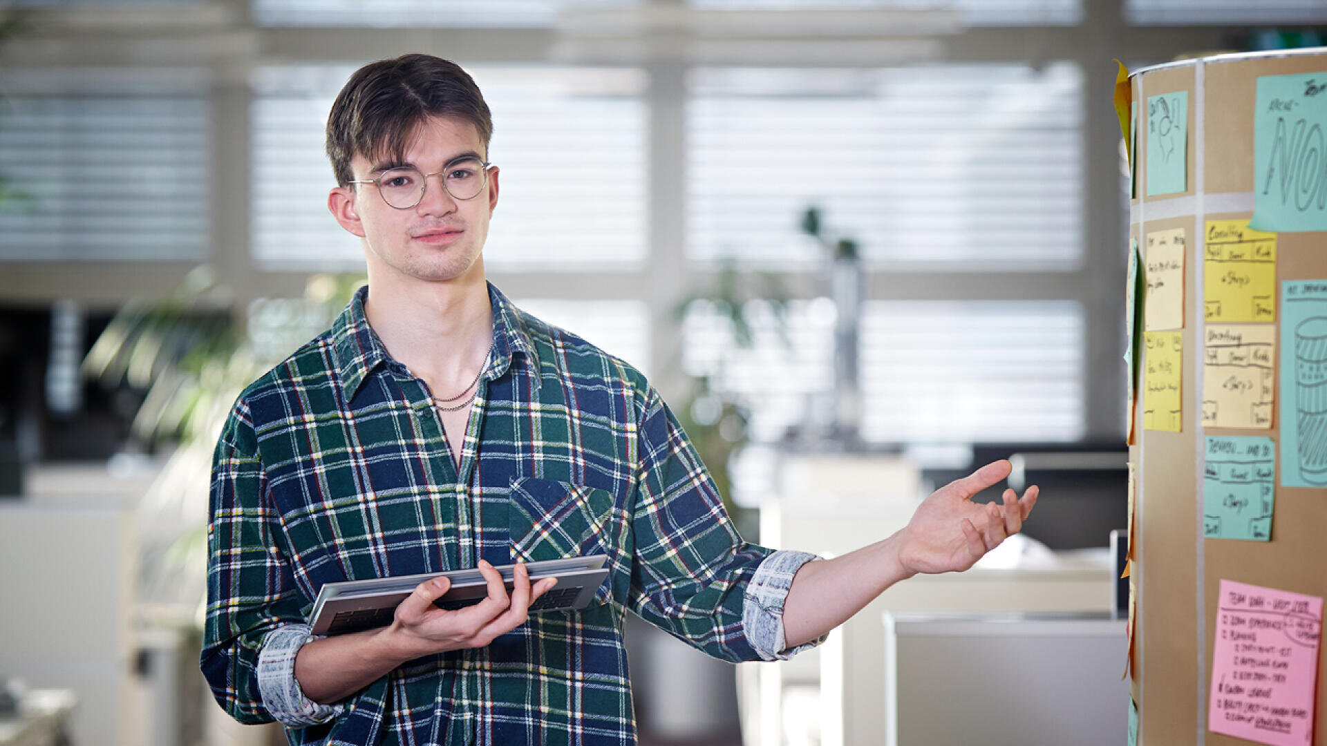 Ein junger Mann mit Brille und grün kariertem Hemd hält ein Tablet in der Hand und deutet mit einer Geste auf eine Tafel mit bunten Haftnotizen in einem hellen, modernen Büro.