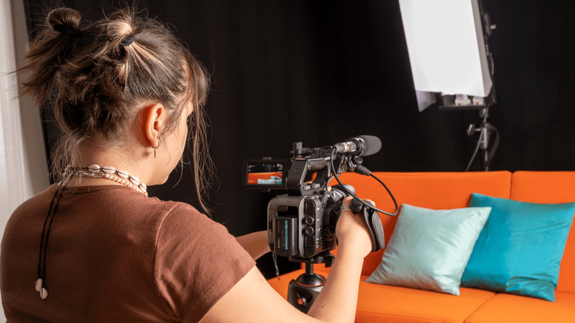 Eine Person mit braunem Haar in einem Dutt filmt eine orangefarbene Couch mit blauen und grünen Kissen mit einer Videokamera in einem Studio mit professioneller Beleuchtung.